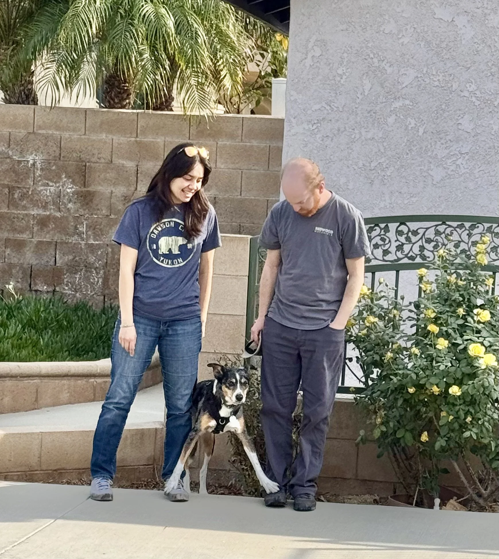 Two people, Asian American woman and Caucasian man, standing outdoors with a puppy dog between them. The woman is smiling while the man is looking down at the dog.