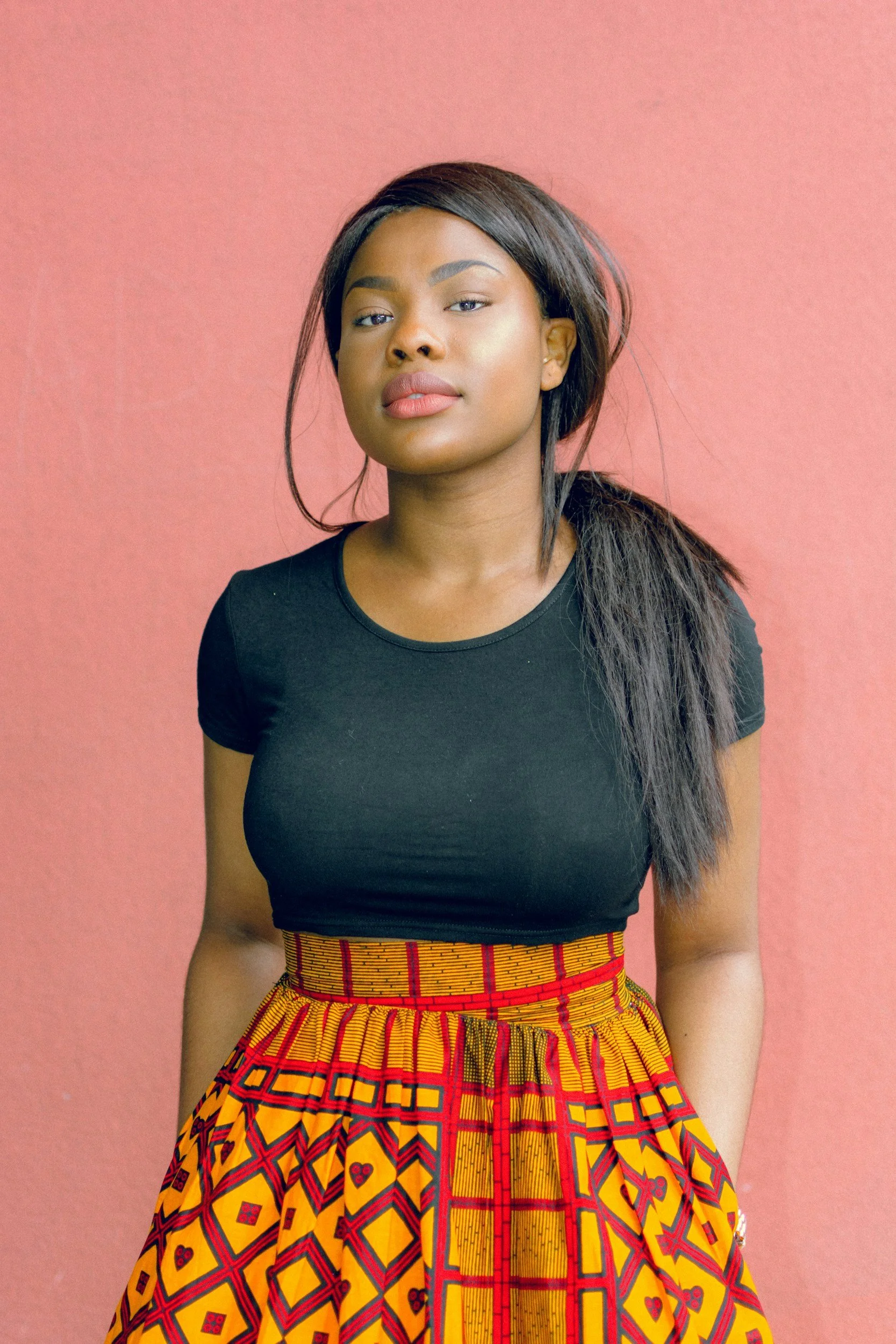 African-American woman with dark hair and a confident expression, wearing a black top and a vibrant, patterned skirt, standing against a pink background.