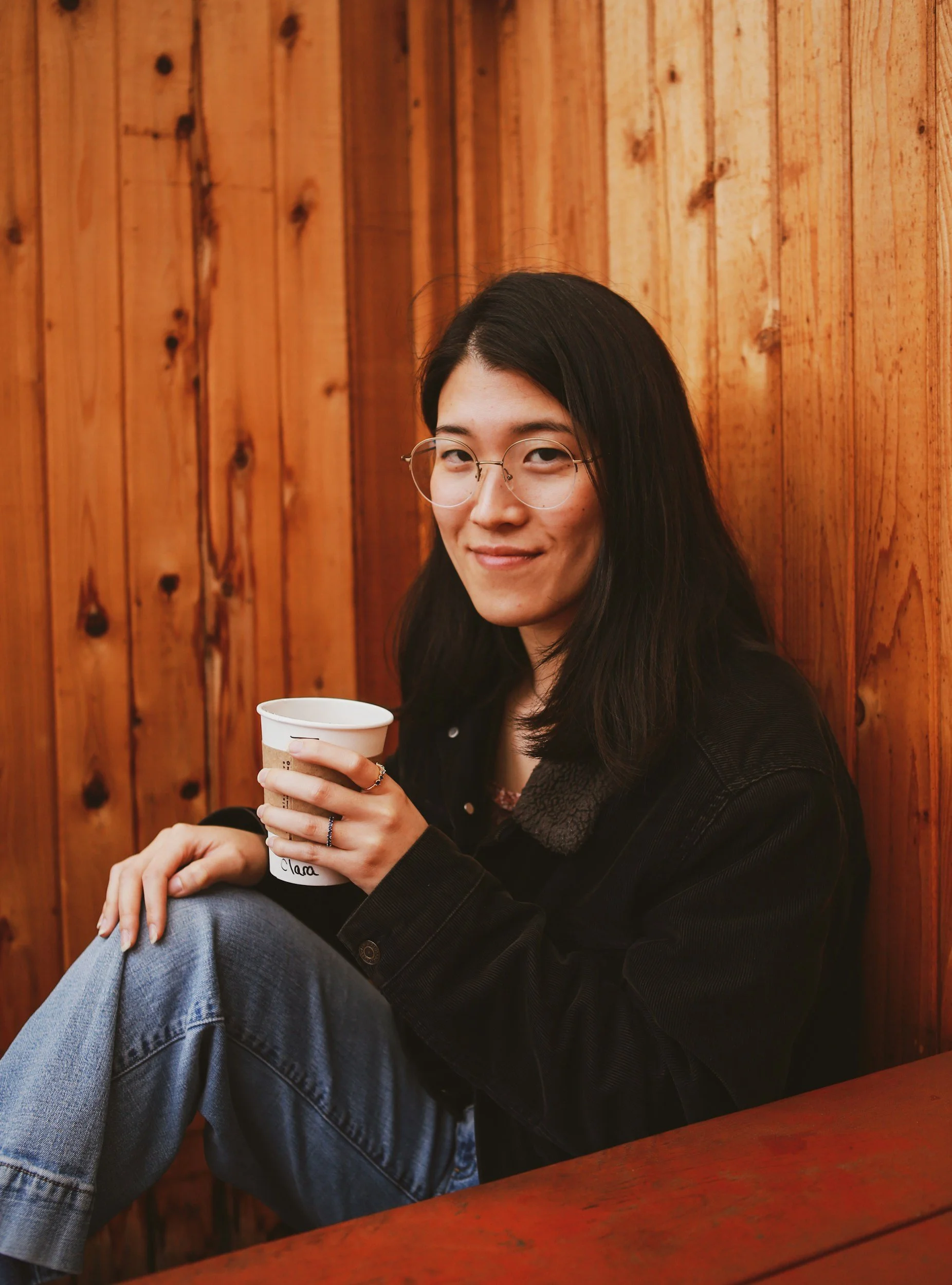 Asian American woman with long black hair, wearing glasses, a black jean jacket with fleece lining, is sitting against a wooden wall with vertical planks and holding a paper cup with her right hand. She is smiling and looking towards the camera.