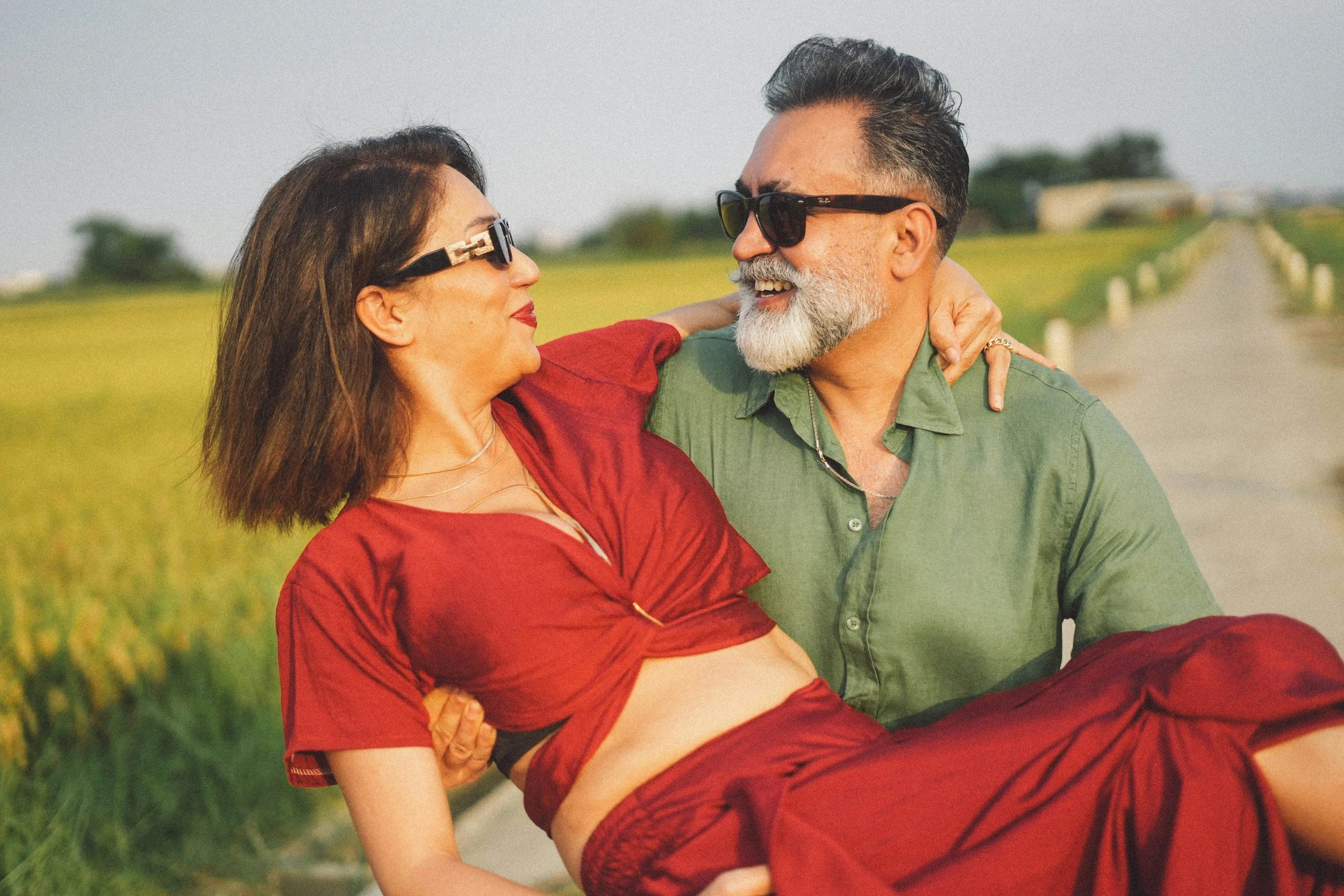 A happy older couple outdoors, embracing and smiling at each other, with a scenic green field and cloudy sky in the background.