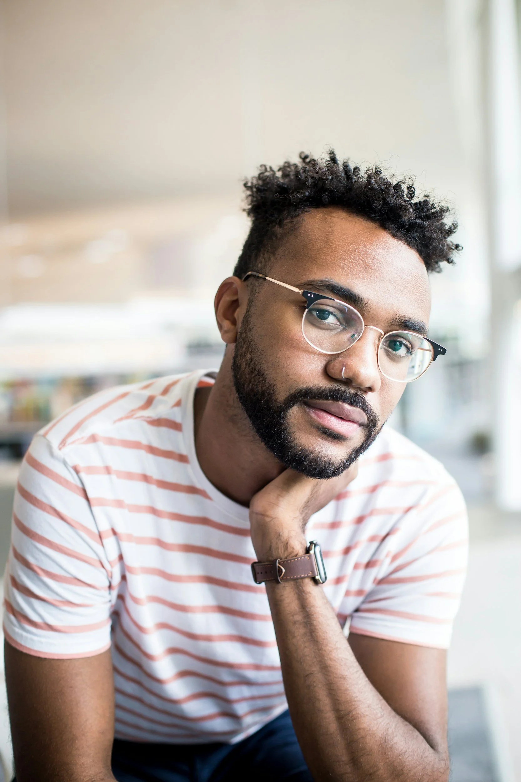 African-American man with glasses, a beard, and curly hair, wearing a white and pink striped shirt, sitting indoors with his chin resting on his hand and looking at the camera.