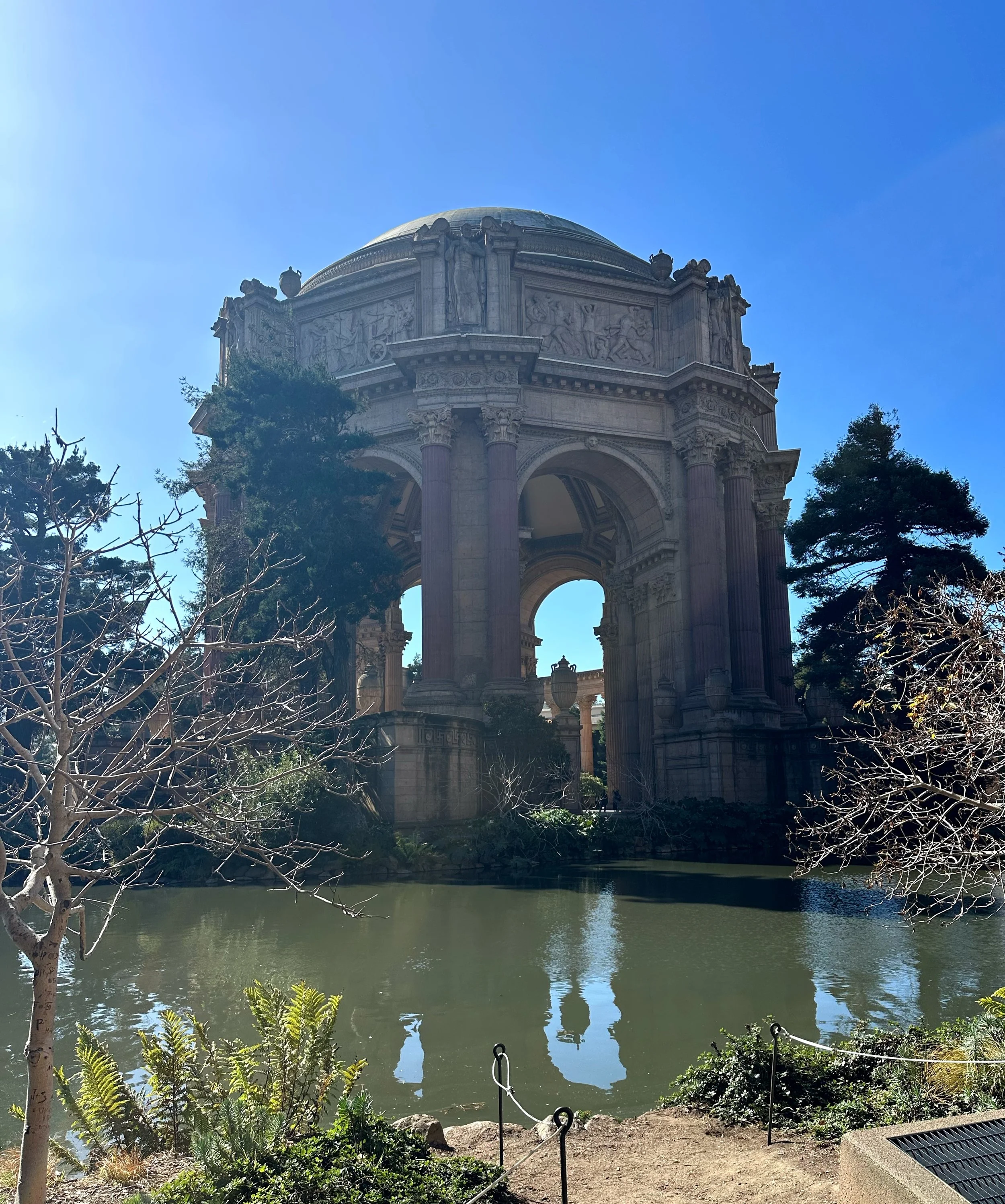 A historic monument with classical architectural design, stone sculptures, and a domed roof, situated beside a reflective pond surrounded by trees and greenery.