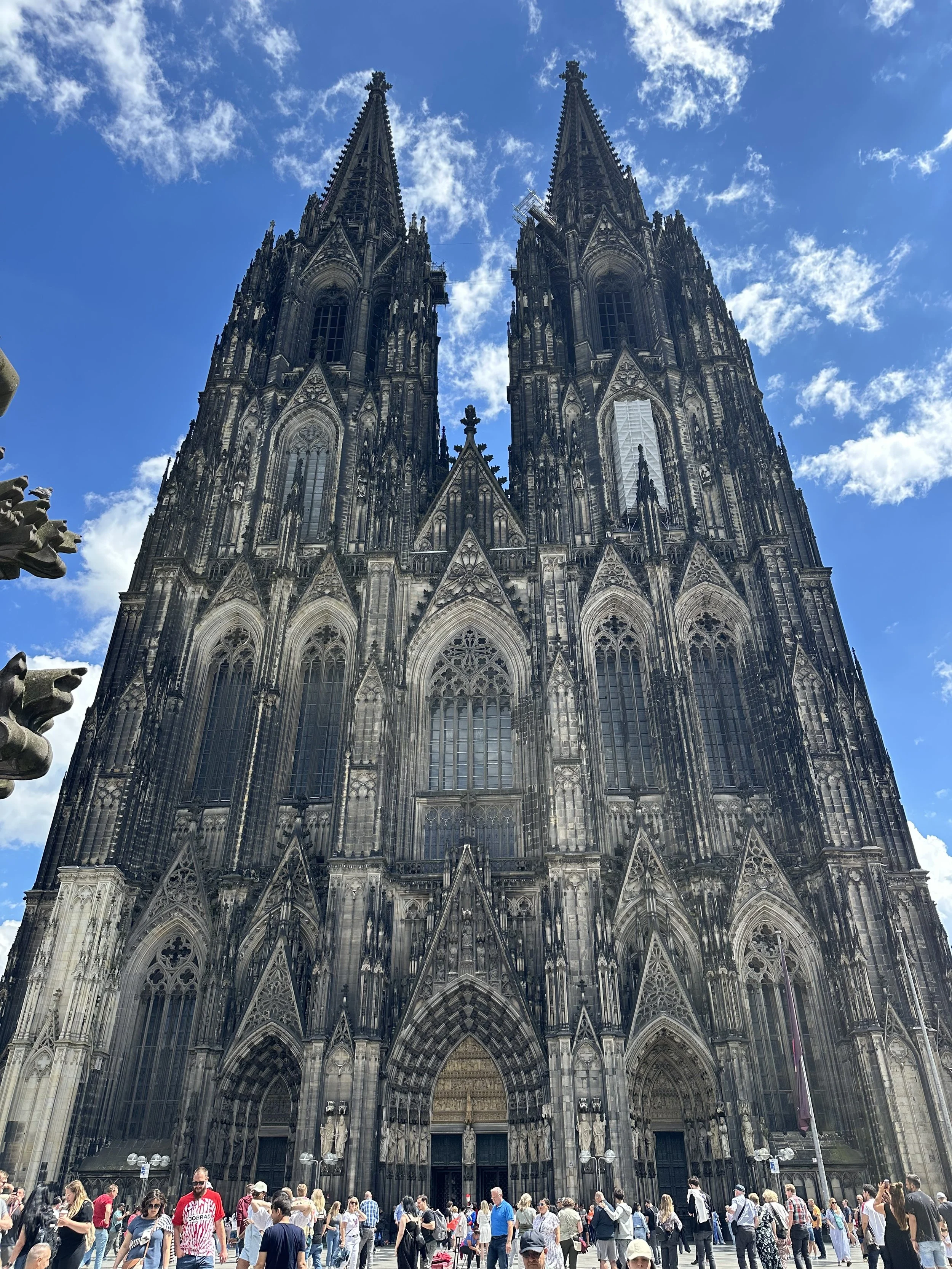 View of Cologne Cathedral with a large crowd of tourists at the entrance on a sunny day with blue sky and some clouds.