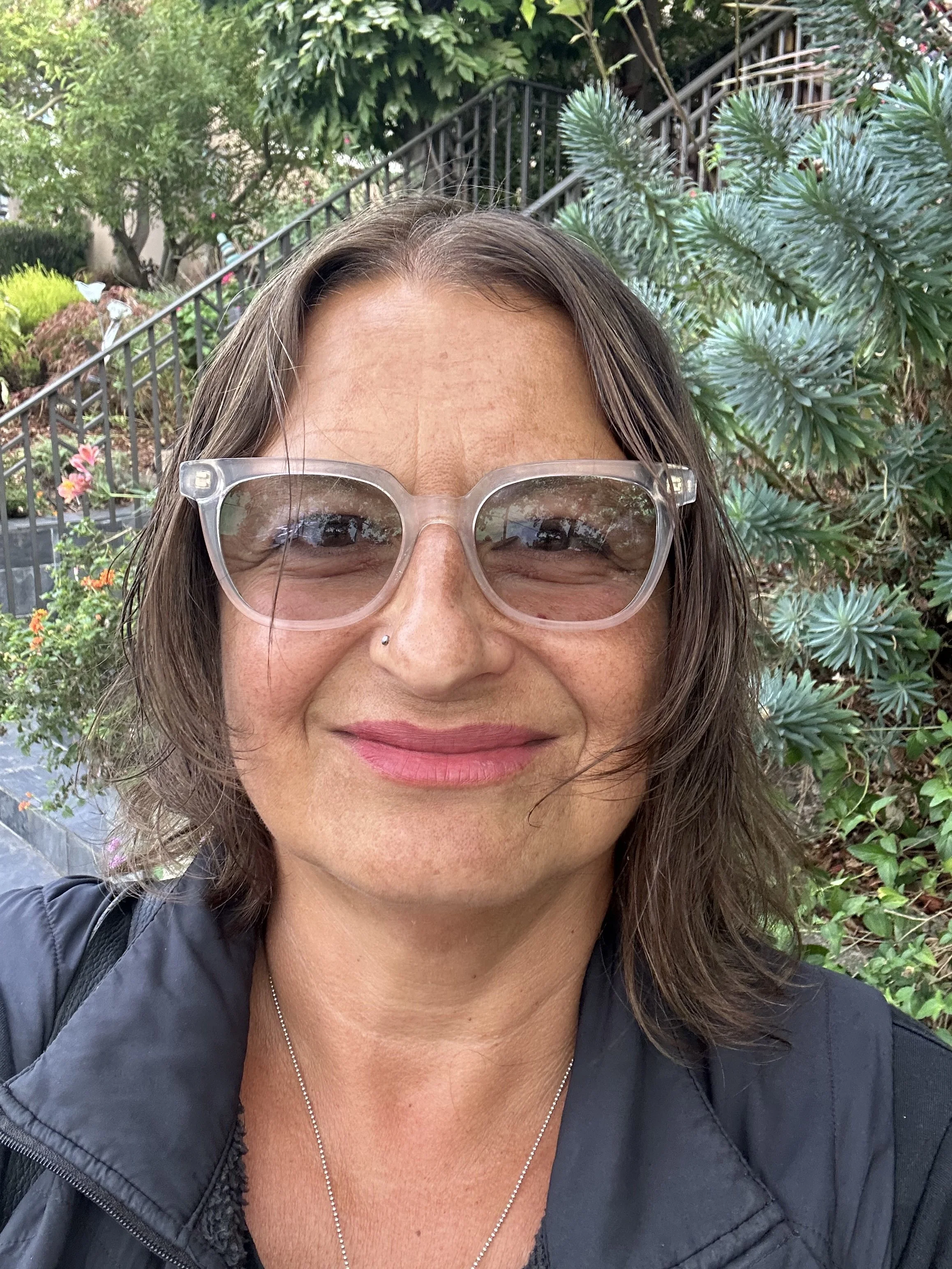 Close-up of a smiling psychotherapist in Oakland, California with wavy brown hair and clear glasses, standing outdoors in a garden with green plants and stairs in the background.