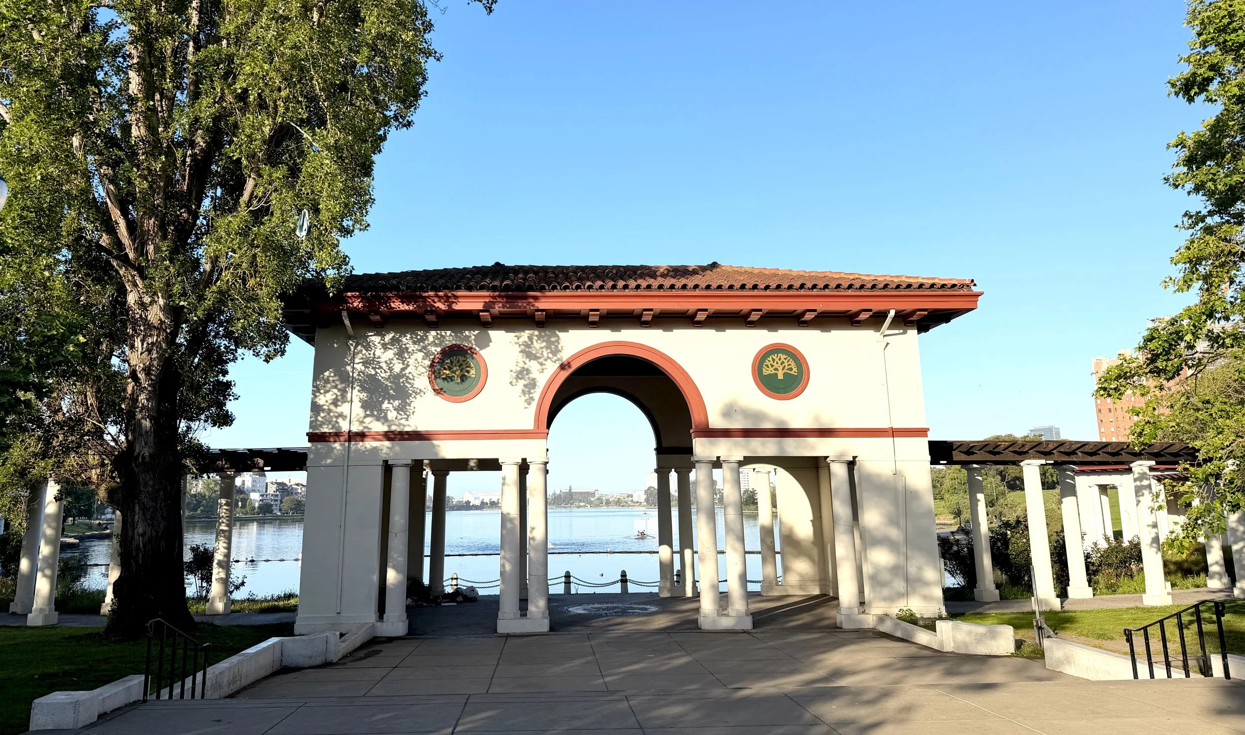 A white pavilion with a red tiled roof and columns, overlooking a river with boats, surrounded by trees, against a clear blue sky.