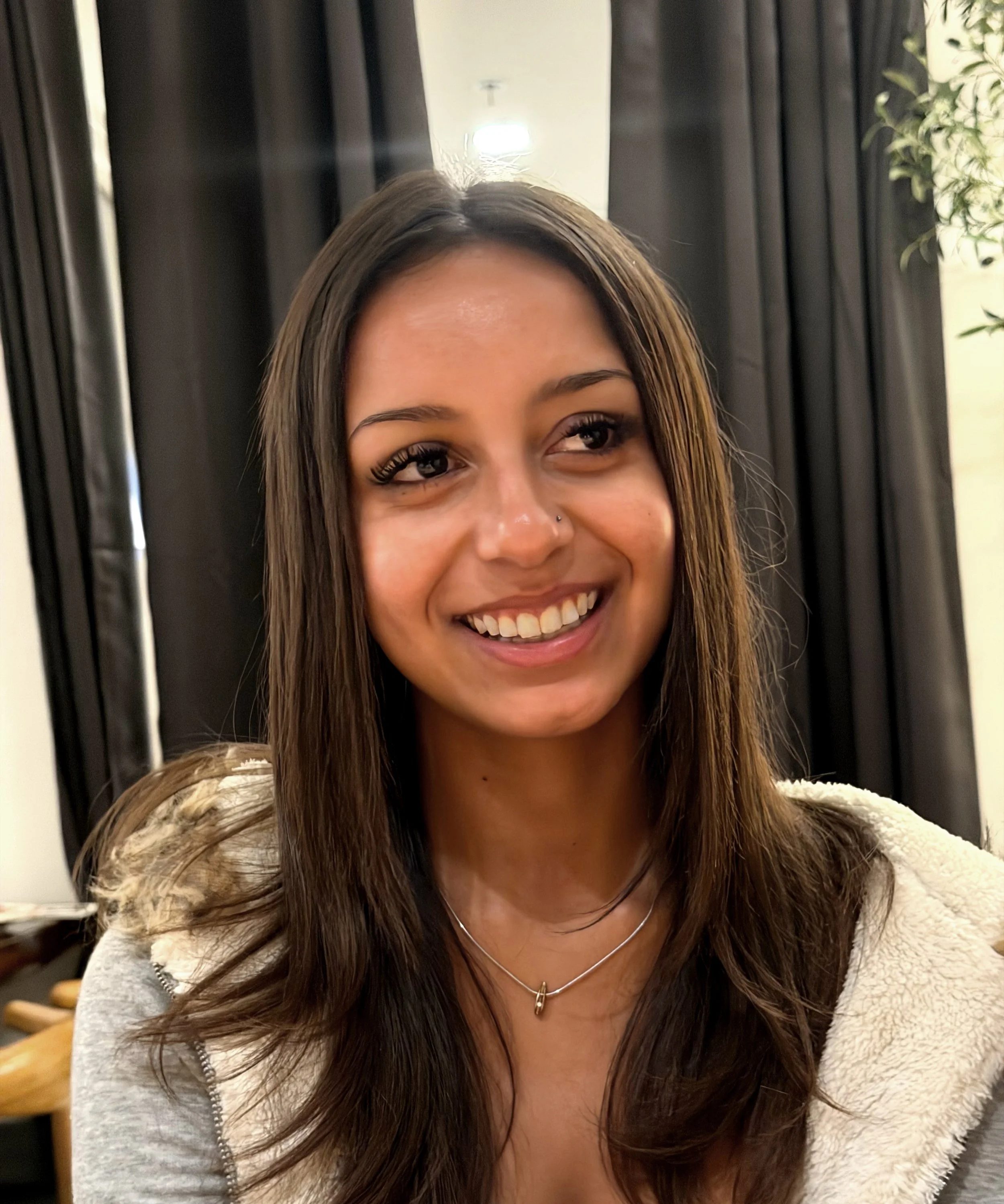 Close-up of a mixed race smiling young woman with long brown hair, wearing a beige sweater, silver necklace, and small facial piercings, sitting indoors.