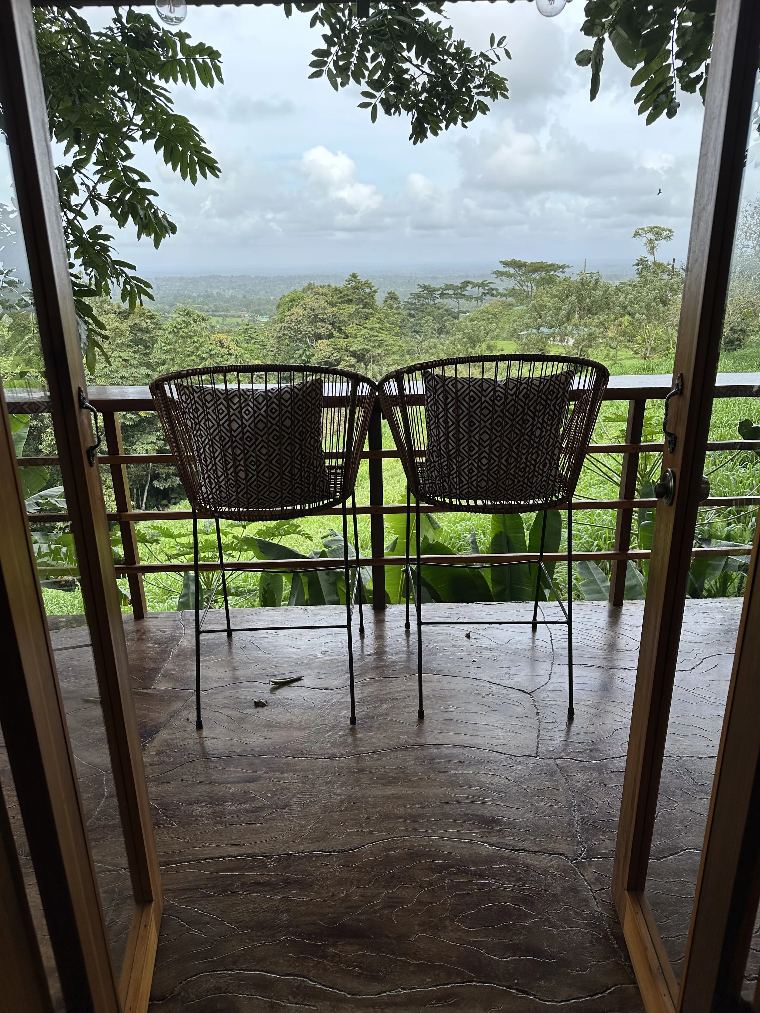 View from a balcony overlooking lush tropical green trees and distant landscape under a cloudy sky, with two empty chairs on a deck in the foreground.