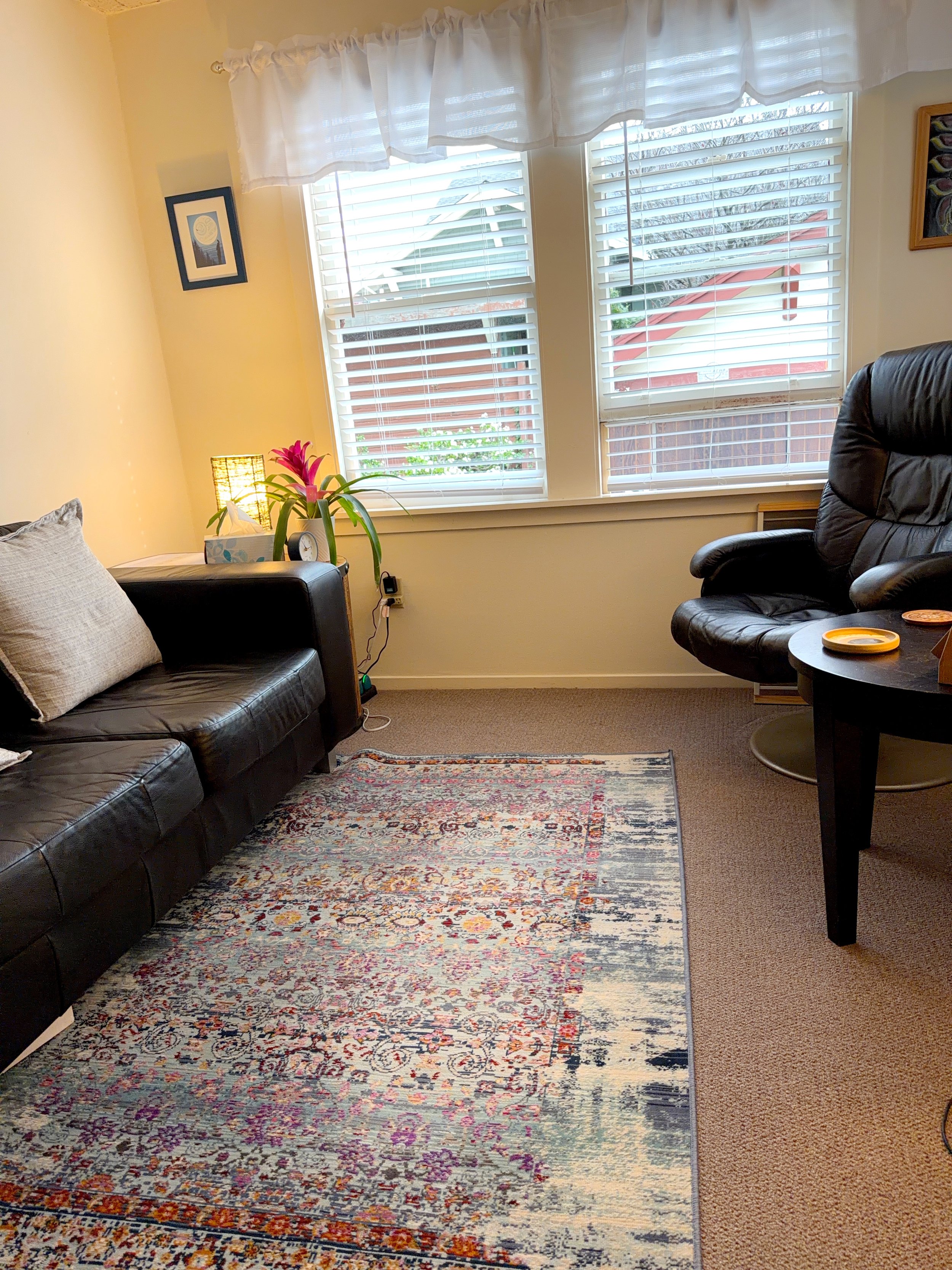 Cozy, warm, inviting psychotherapy room with black leather couch, a colorful patterned area rug, a small side table with a plant and clock, a beige armchair, a window with white blinds and curtains, and framed artwork on the yellow walls.