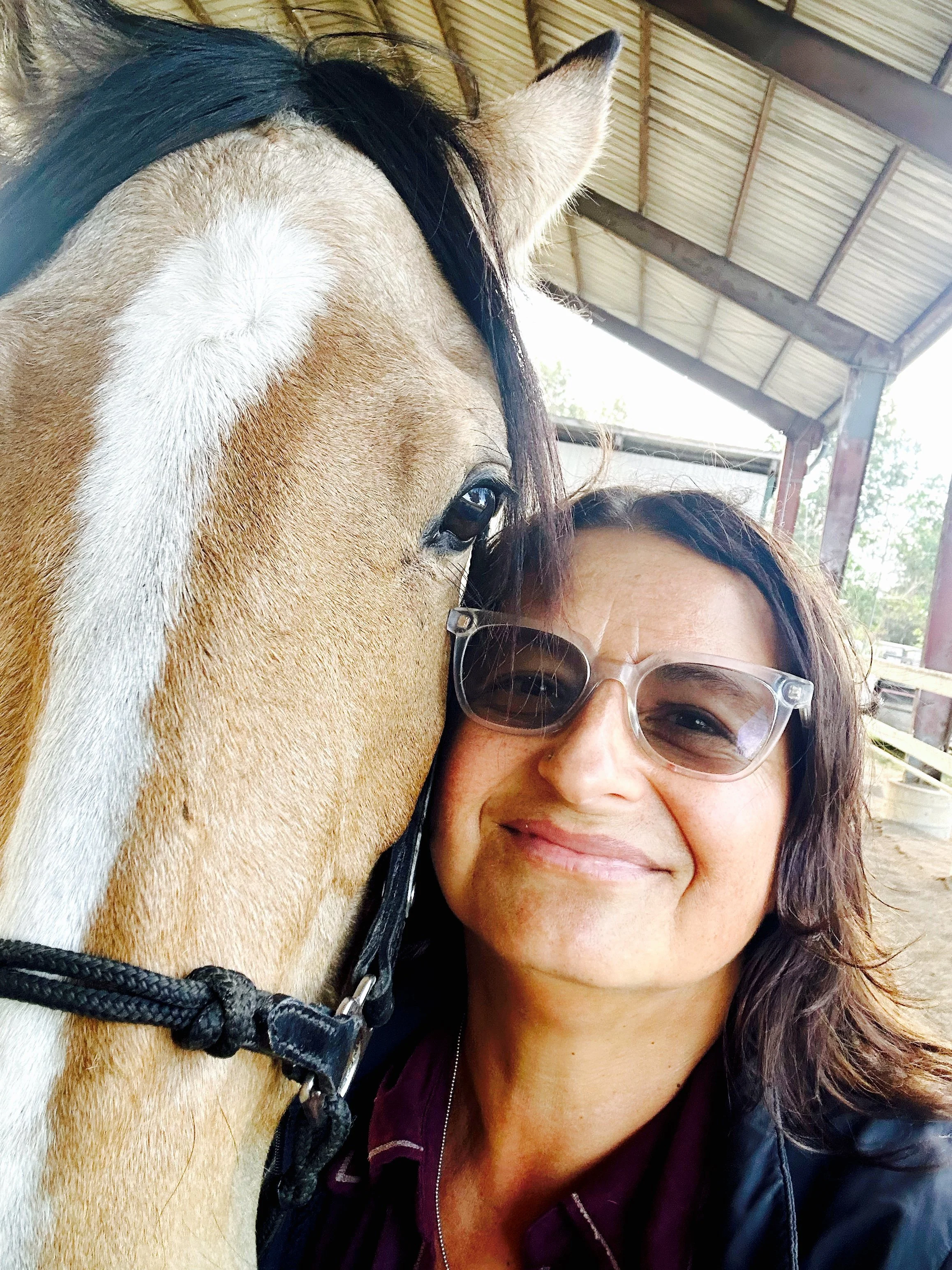 Female psychotherapist with wavy brown hair, glasses, and a smile taking a selfie with a tan colored horse with a black mane. Specializes in relationship counseilng, trauma, cultural/racial identity, and anxiety.