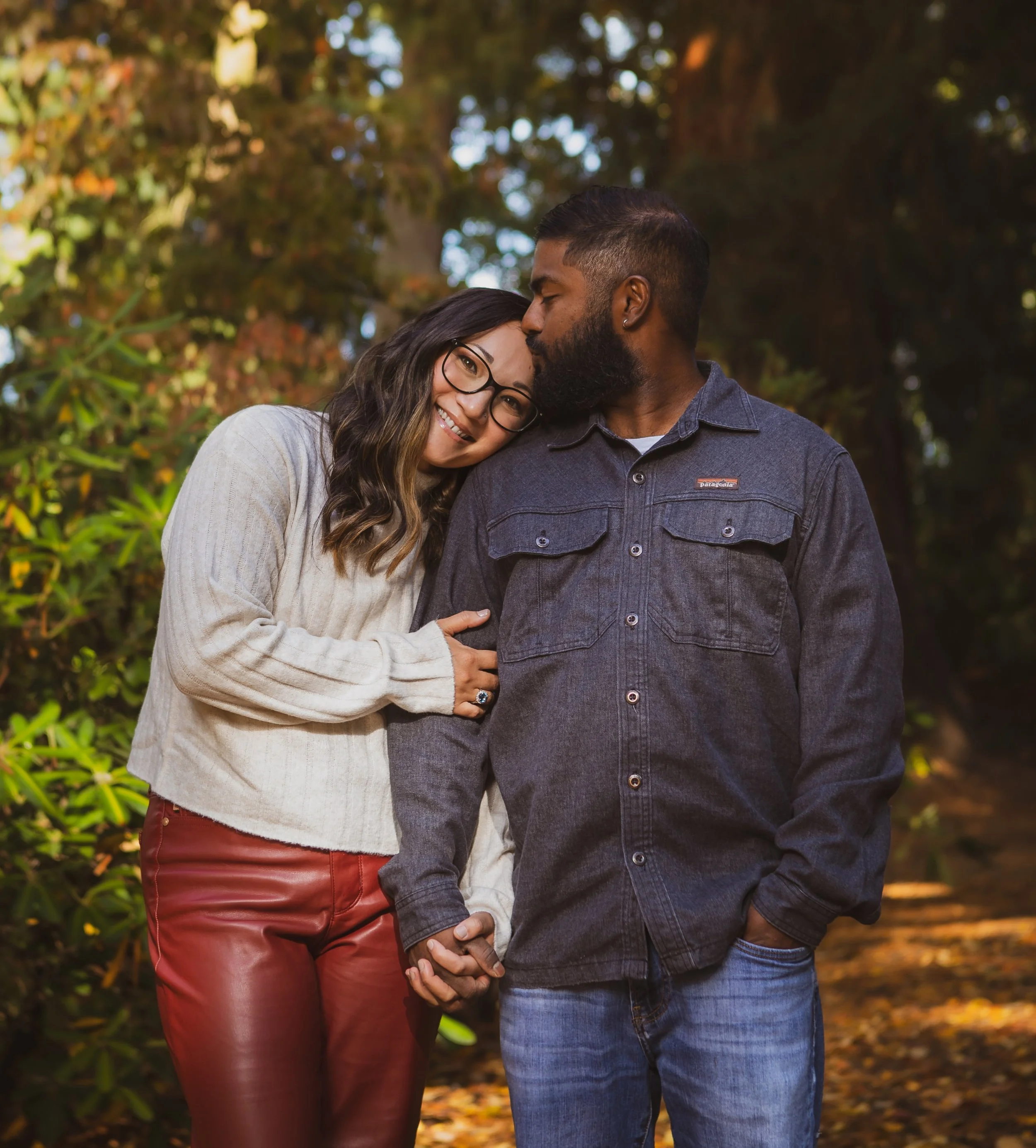 Bi-racial couple holding hands and sharing a kiss, standing outdoors in a park with autumn foliage in the background.