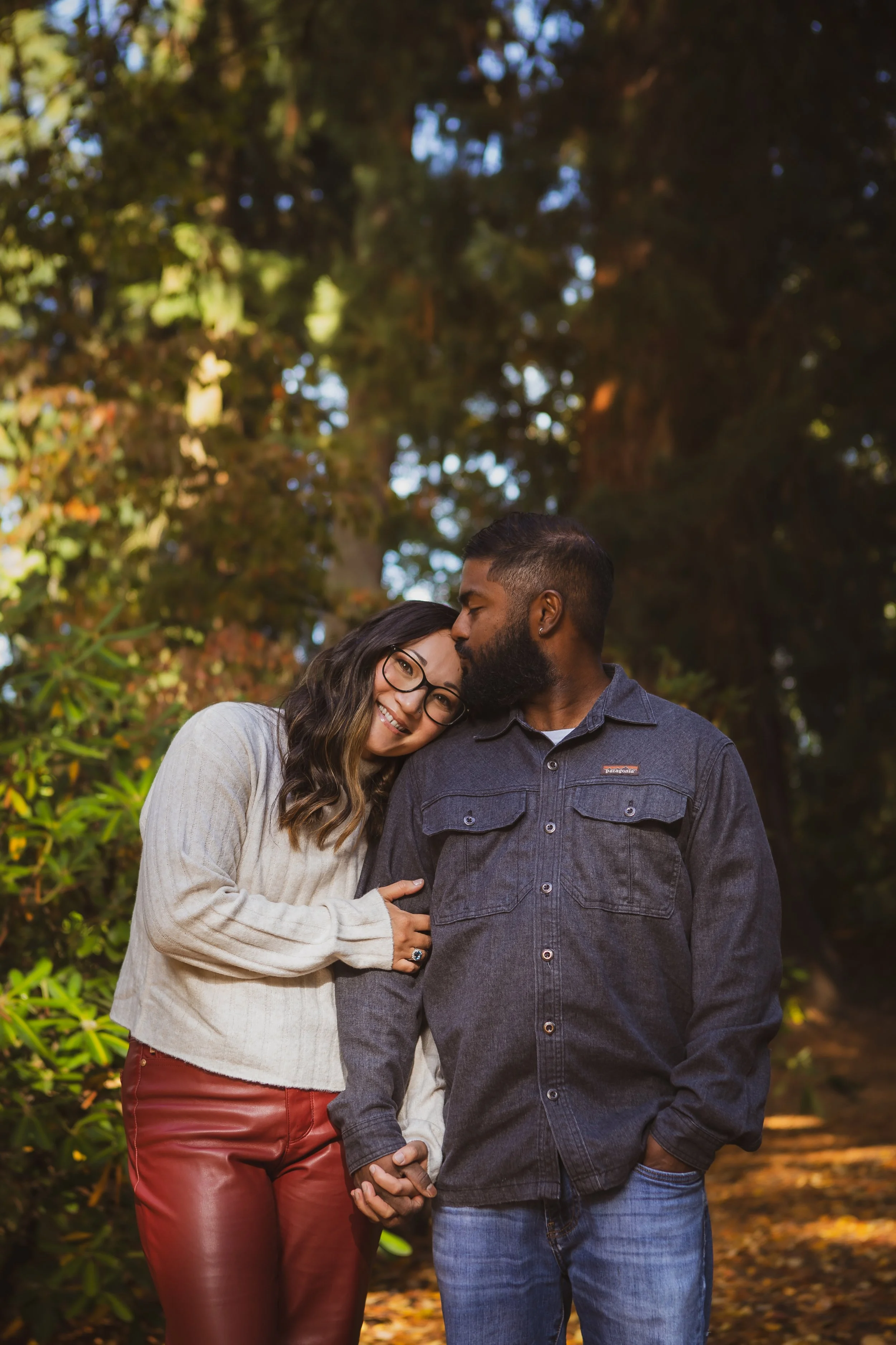 Mxed race couple stands close together outdoors during fall, holding hands and smiling, with trees and autumn leaves in the background.