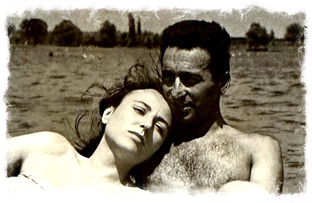 Black-and-white photo of a man and woman close together outdoors, the woman is resting on the man's chest. Trees and water in the background.