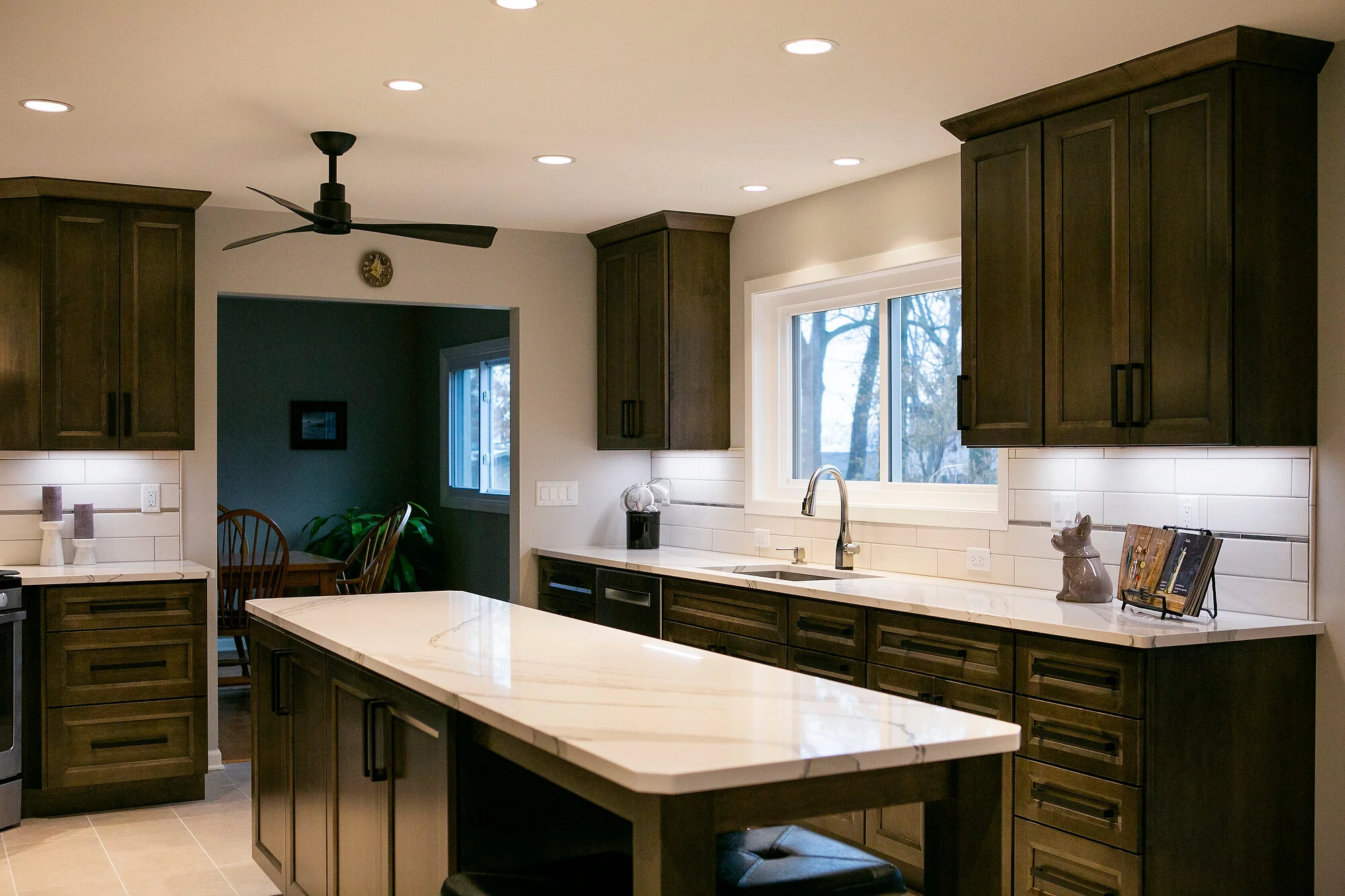  Kitchen with dark brown cabinets, white marble countertops, black ceiling fan, and central island. 