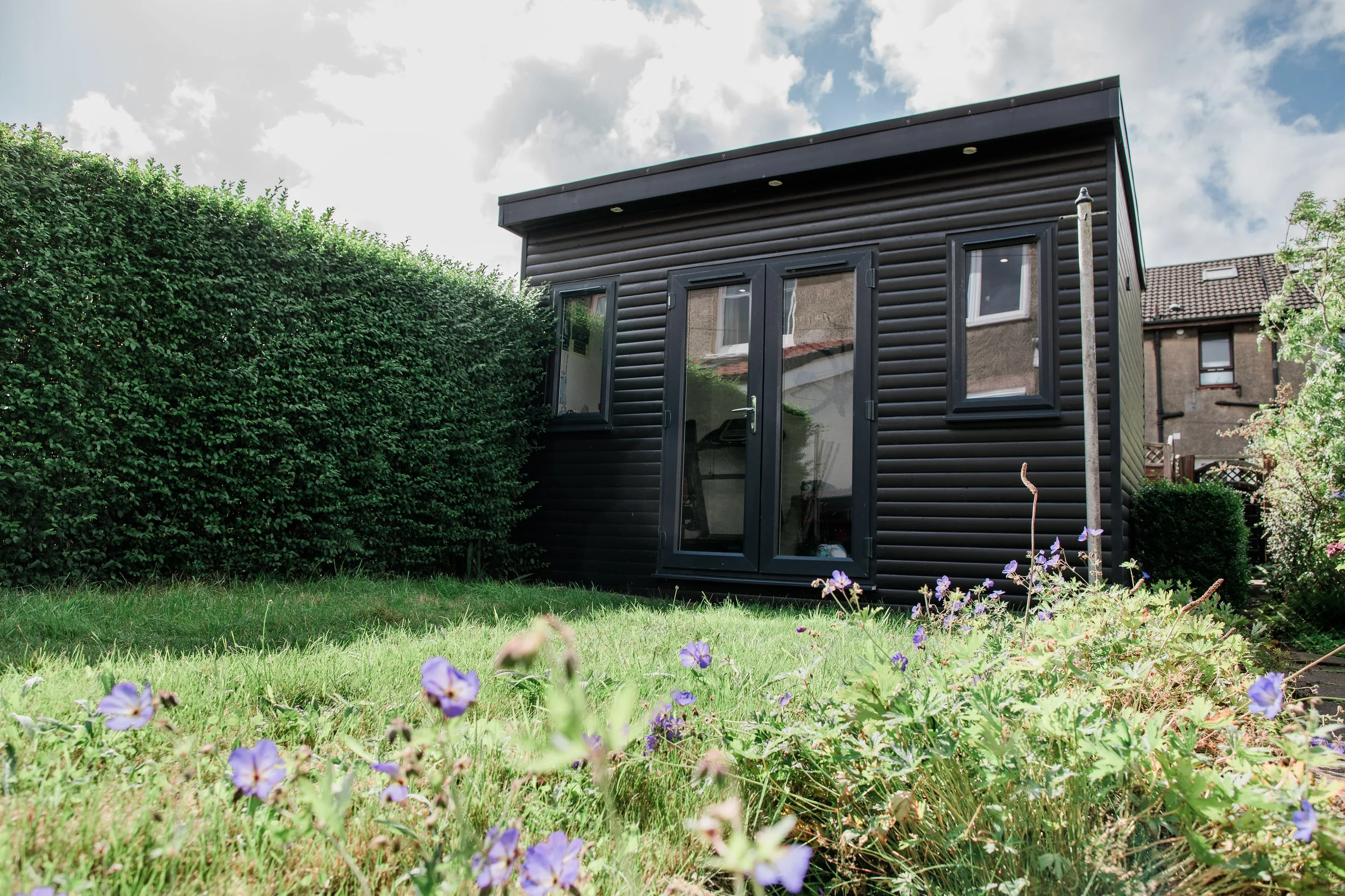 Black garden shed with a double glass door and two windows, surrounded by greenery and purple flowers in a backyard.