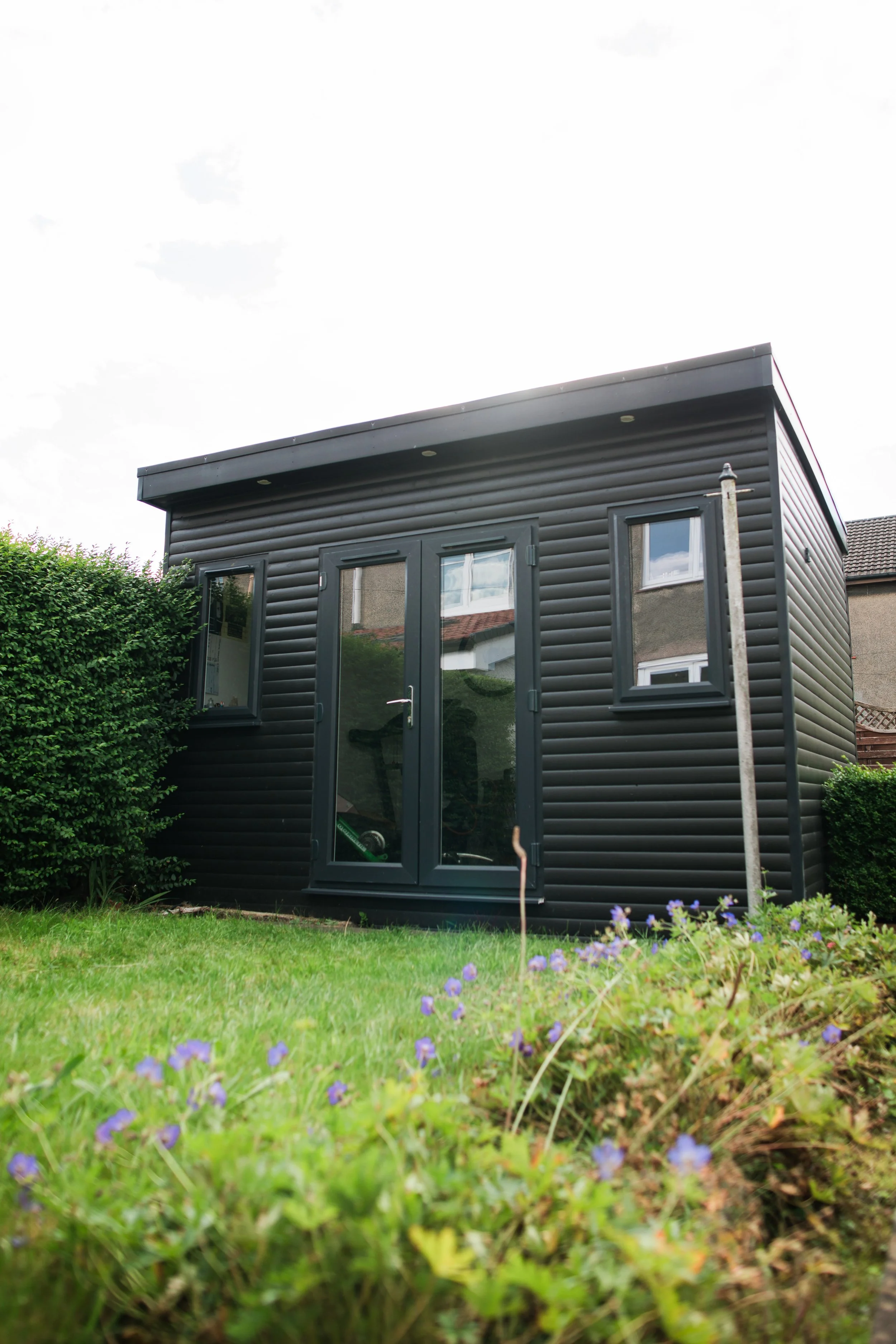 A small black modern shed with double glass doors and two windows on a grassy lawn, surrounded by bushes and flowers.