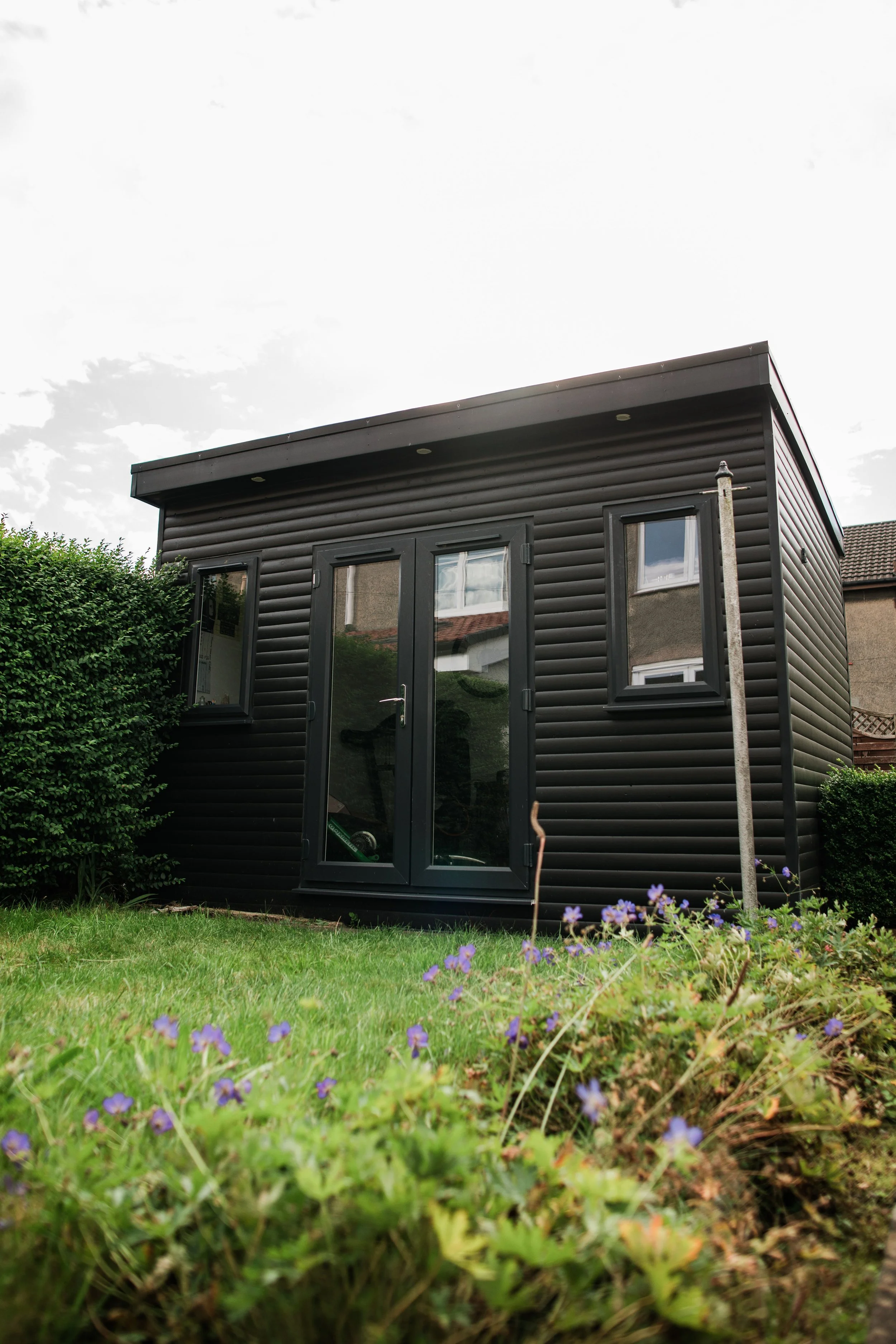 A small black garden shed with double glass doors, surrounded by greenery and purple flowers in the foreground.