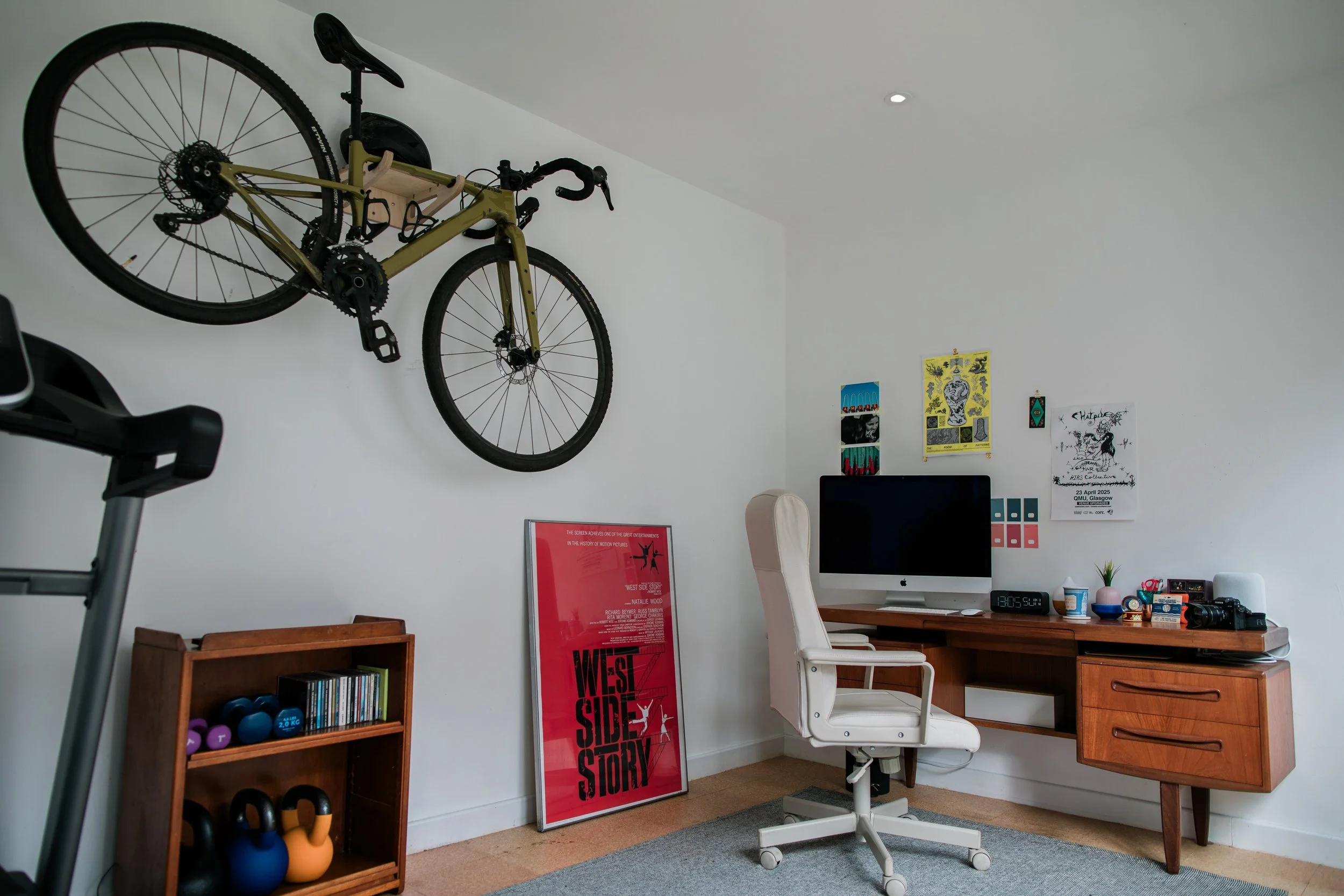 A home office with a wall-mounted bicycle, a wooden bookshelf with exercise weights, CDs, and kettlebells, a framed "West Side Story" poster, a desk with a computer, various decorations, and an ergonomic white office chair.