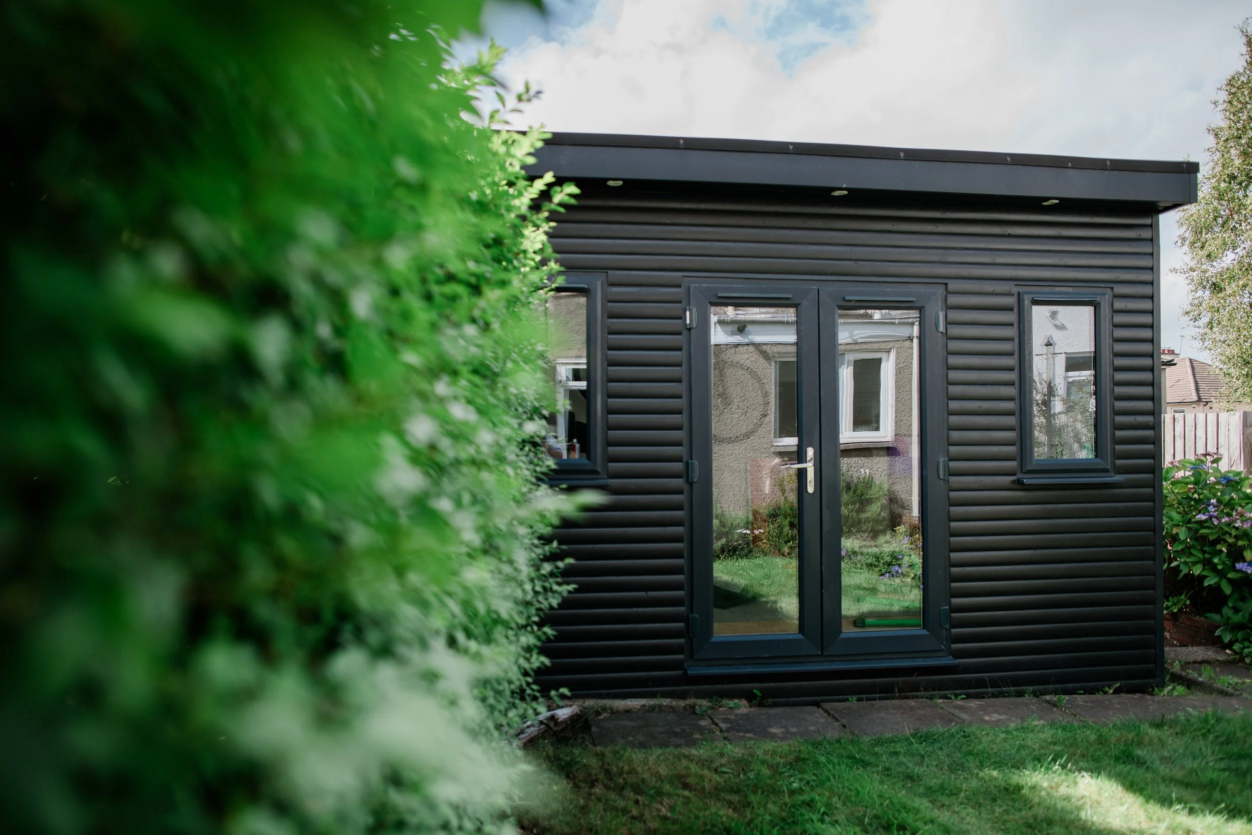 Black garden shed with glass double doors and two small windows, surrounded by greenery and a lawn.