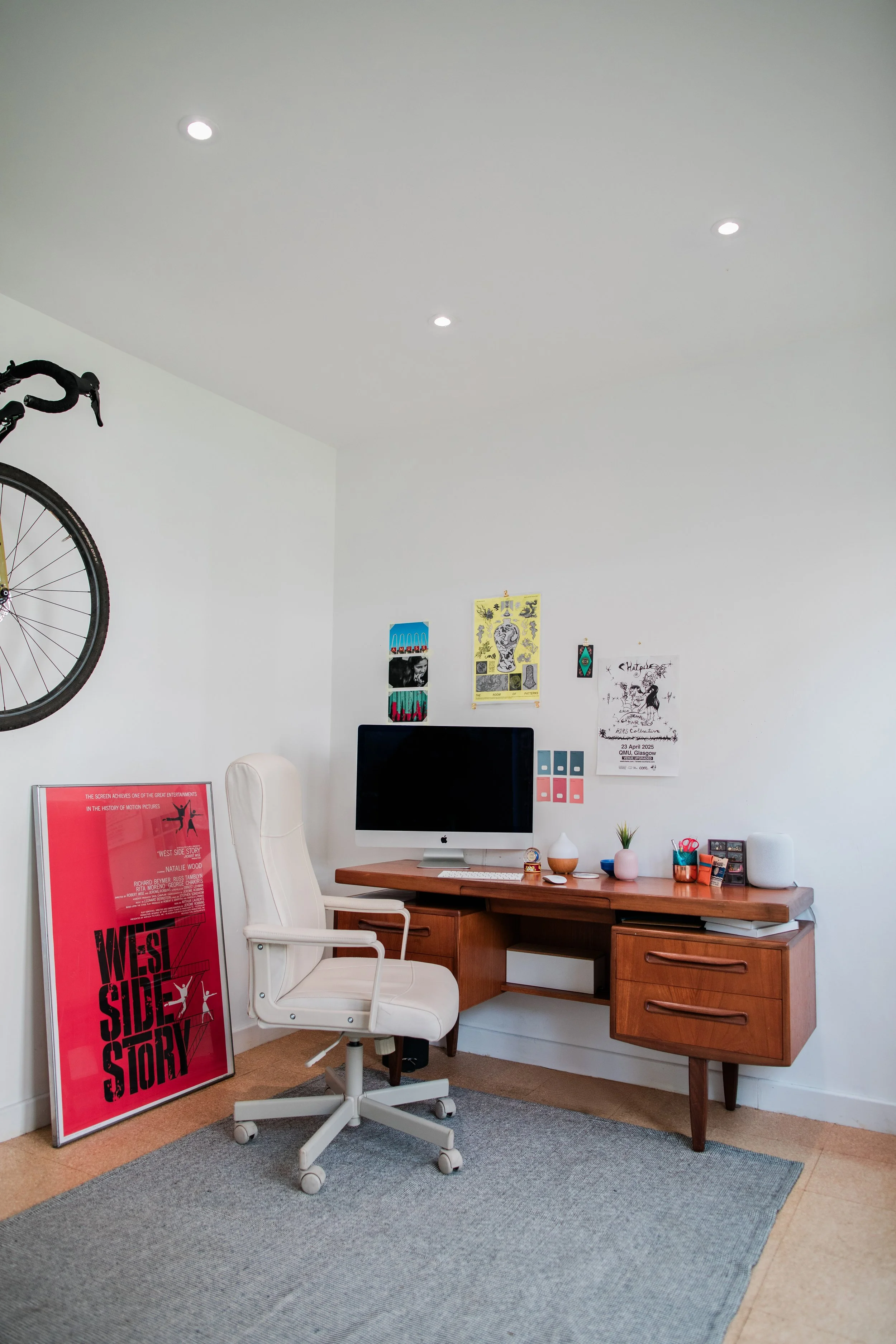 Home office with a wooden desk, white office chair, desktop computer, a framed "West Side Story" poster, bicycle mounted on the wall, and decorative items on the desk, including plants and stationery.