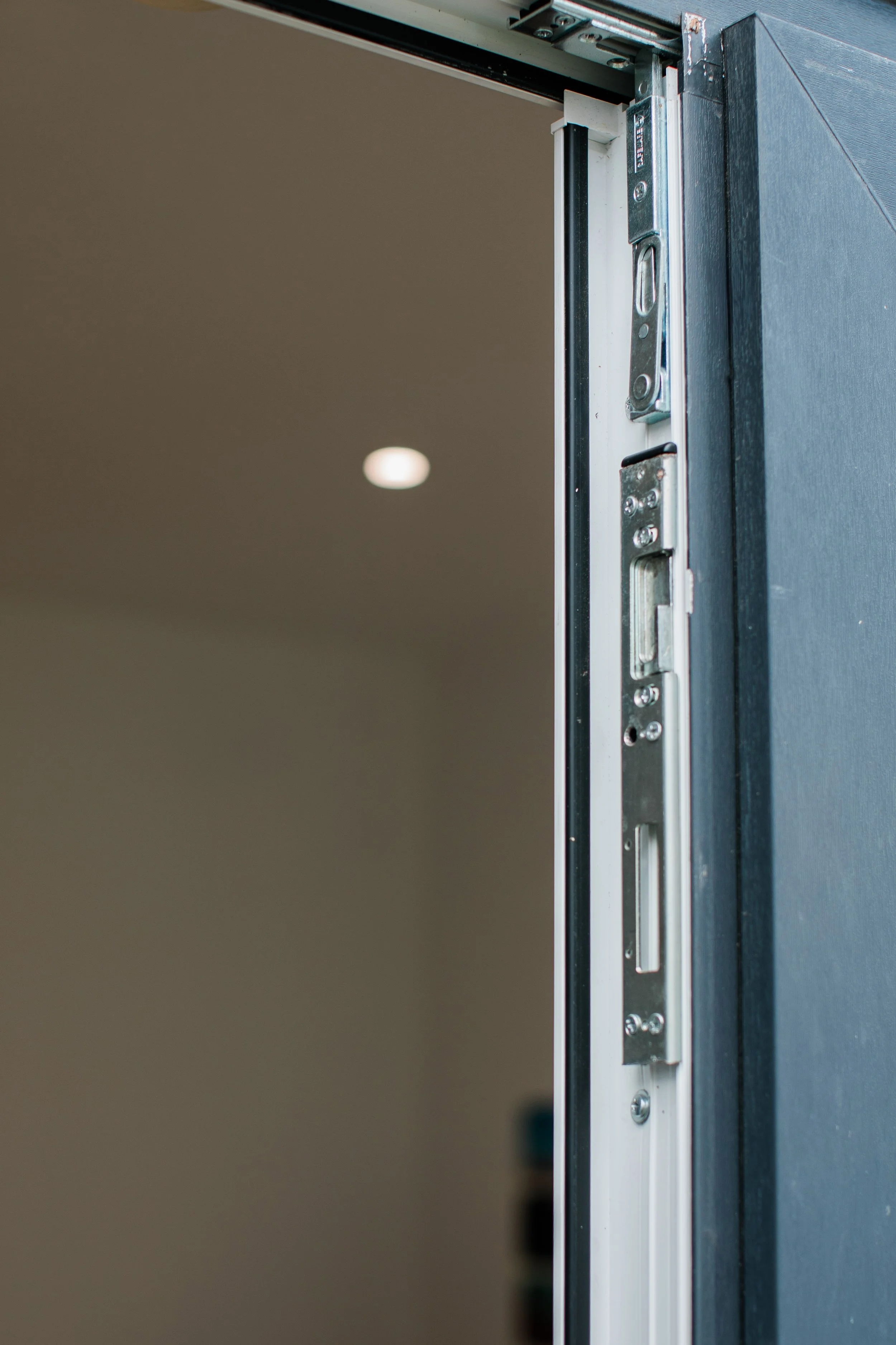 Close-up of a door frame with a metal latch and hardware, with a blurred interior wall and ceiling light in the background.