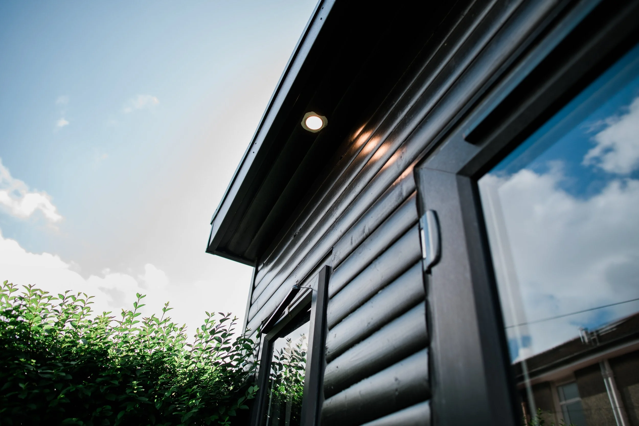Close-up of a modern house exterior with black horizontal siding, a small window, and a single outdoor light fixture under a partly cloudy sky.