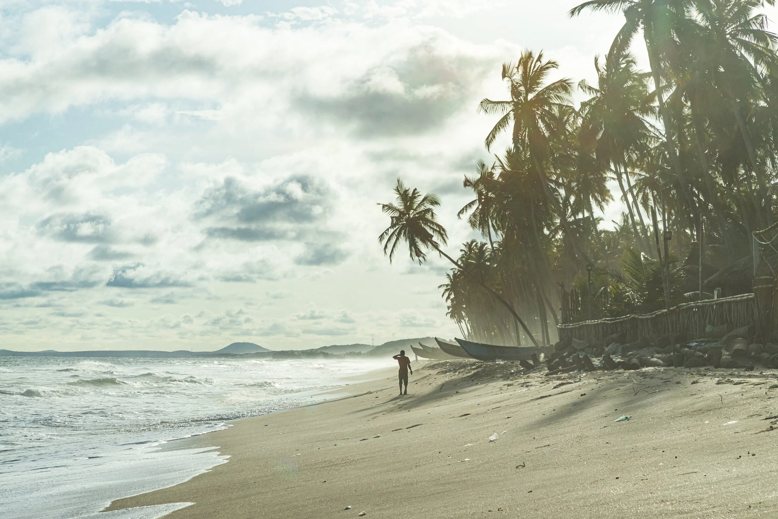 A person standing on a sandy beach near the ocean, lined with tall palm trees and small boats, under a partly cloudy sky.