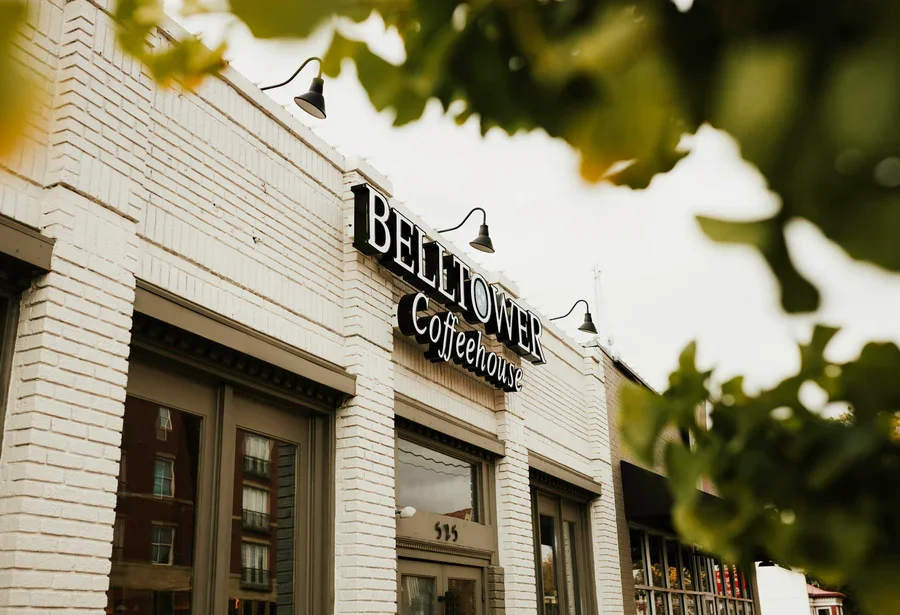 Exterior view of Bellflower Coffeehouse building with white brick facade, black sign, and outdoor lighting, partially framed by green leaves.