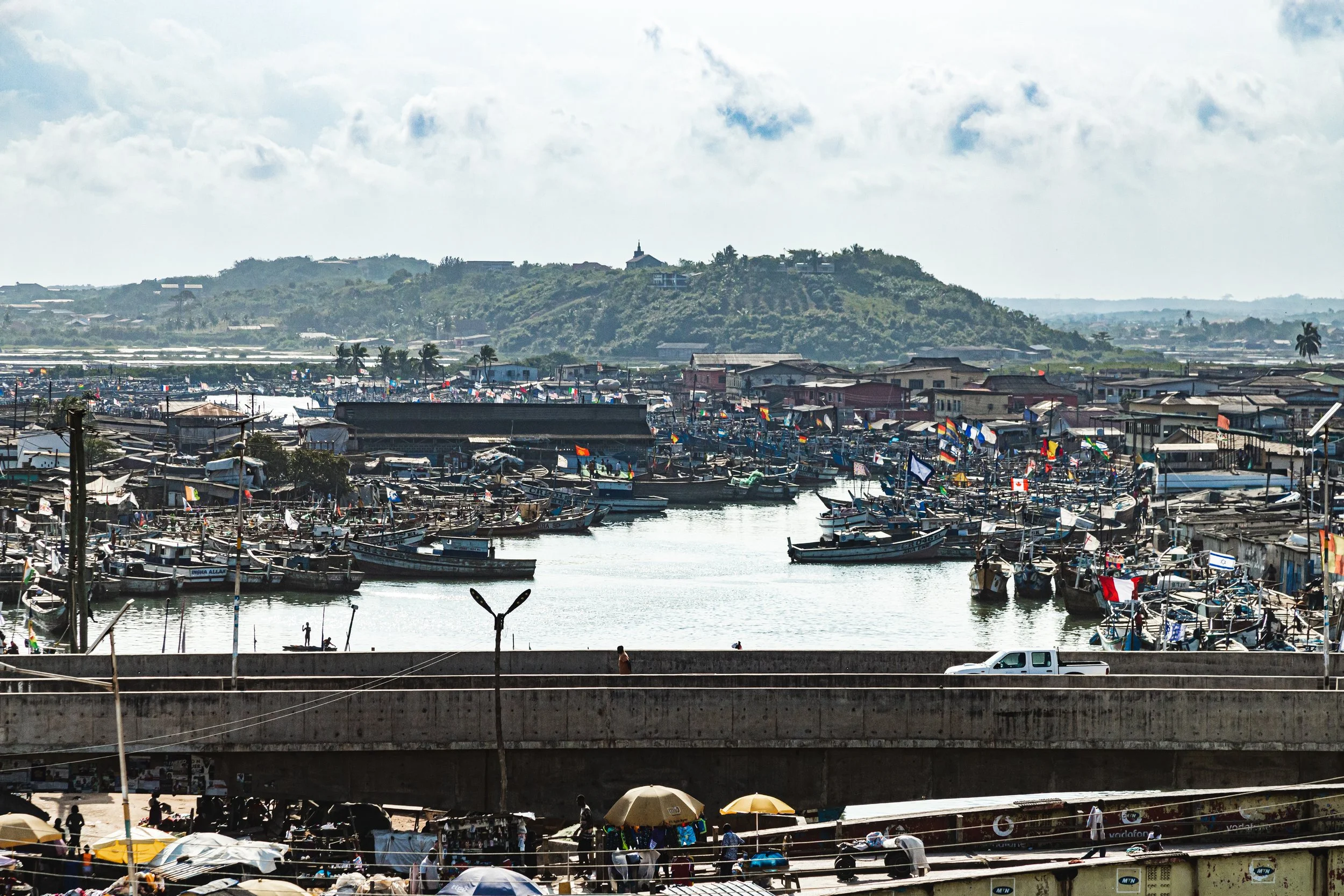 A busy harbor with numerous boats docked along the shoreline, colorful flags fluttering, and a mountain in the background under a partly cloudy sky. People are walking, with some umbrellas providing shade.