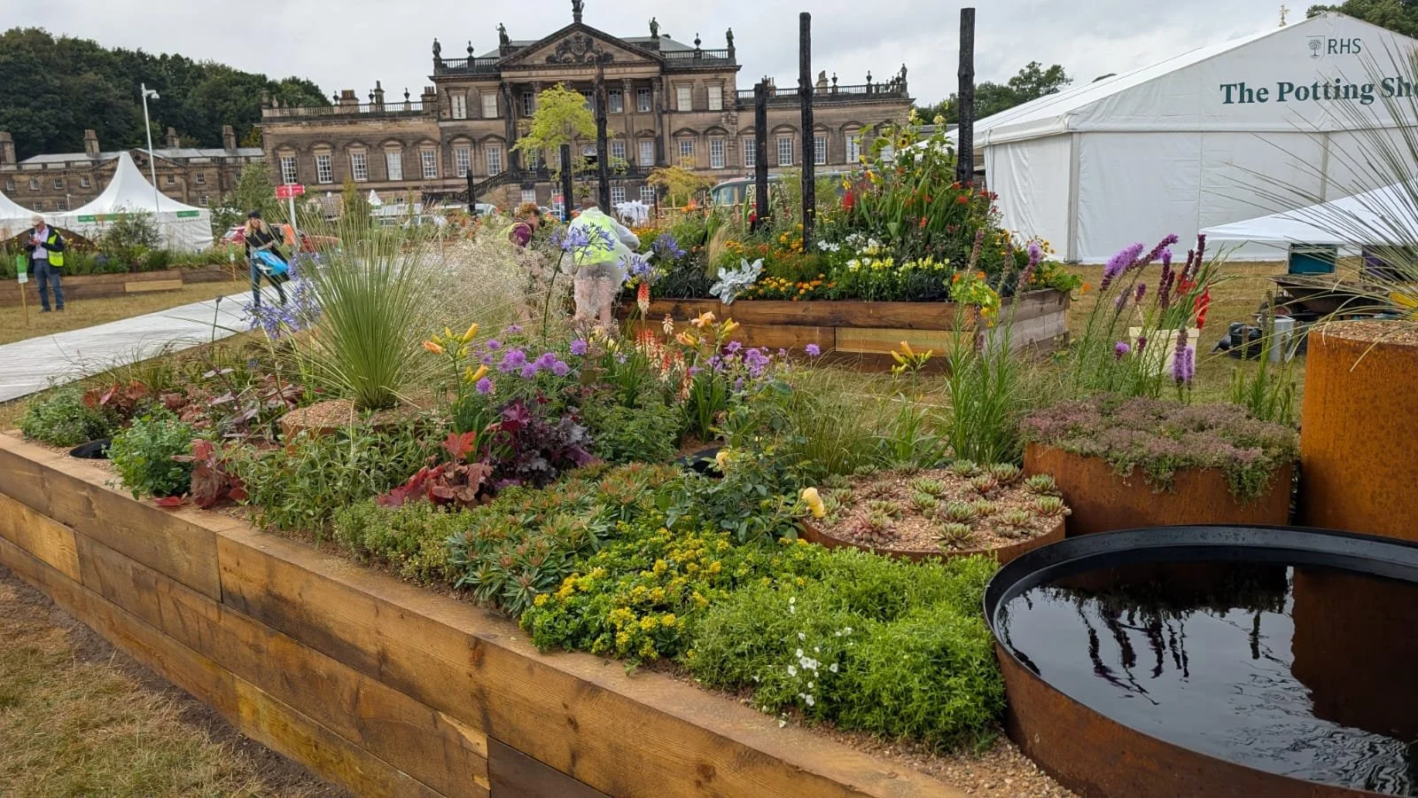View of a garden display with various plants, flowers, and rustic containers, set up at an outdoor event with a historic building and tents in the background.