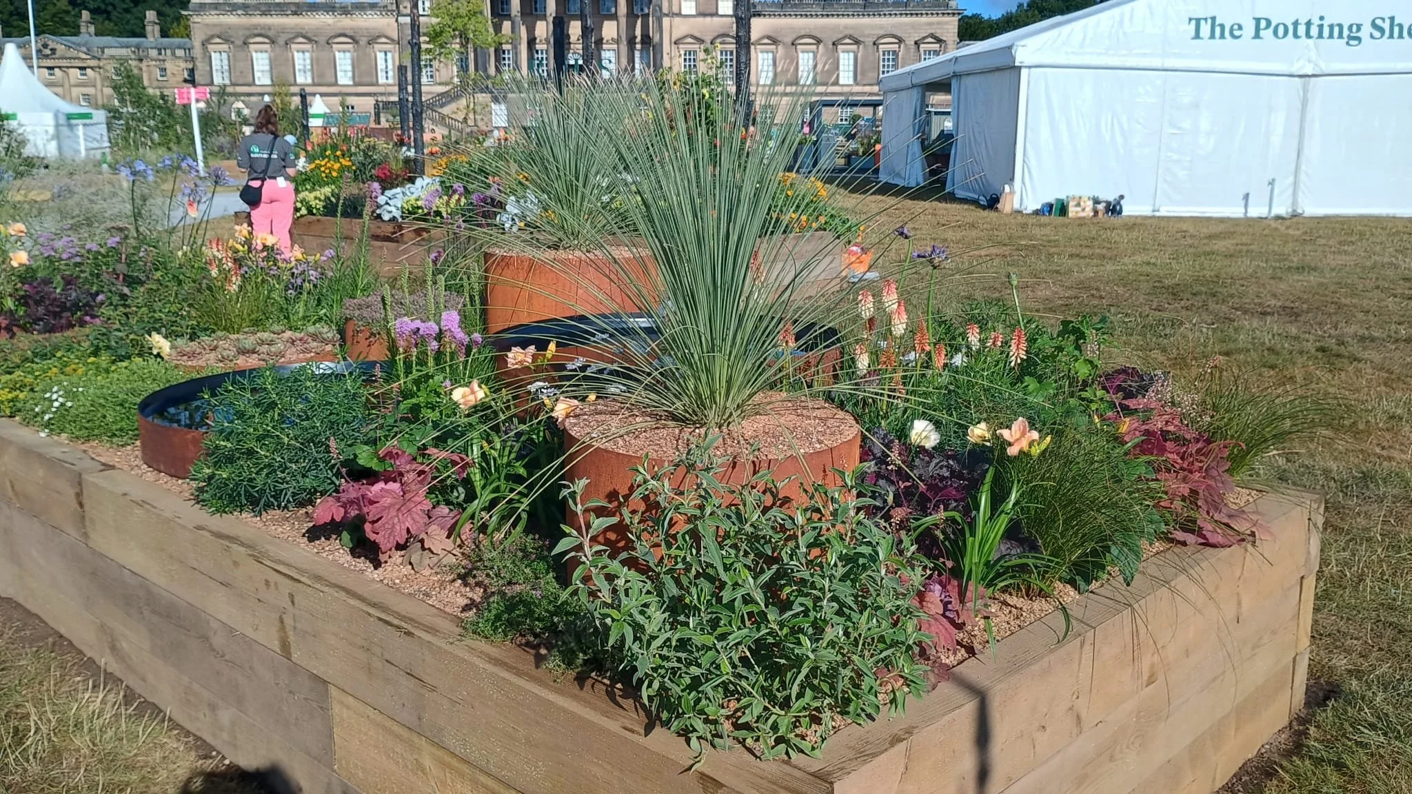 A raised wooden garden bed with a variety of plants and flowers, including tall grassy plants and colorful blooms, at an outdoor event with tents and people in the background.