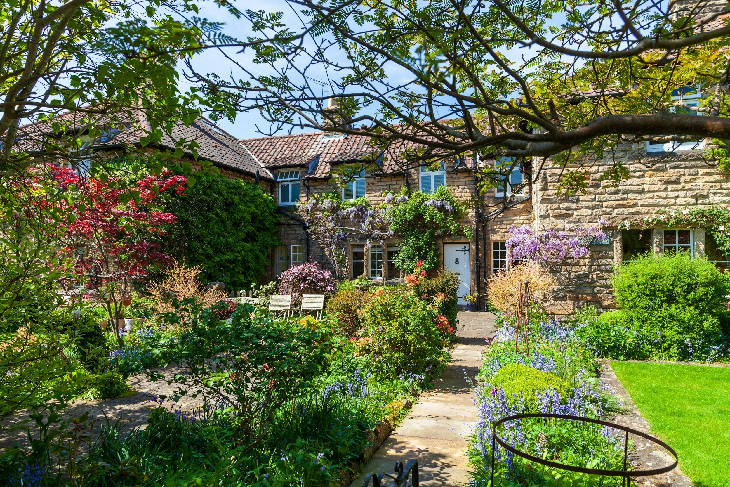 A lush garden pathway lined with colorful flowers and green shrubs on both sides, with a brick wall on the left and a variety of trees in the background.