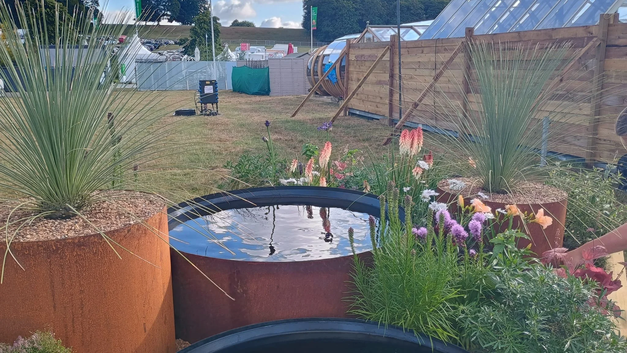 Plants in large rust-colored containers, including grasses and flowering plants, with a small water feature, in an outdoor garden or park setting.