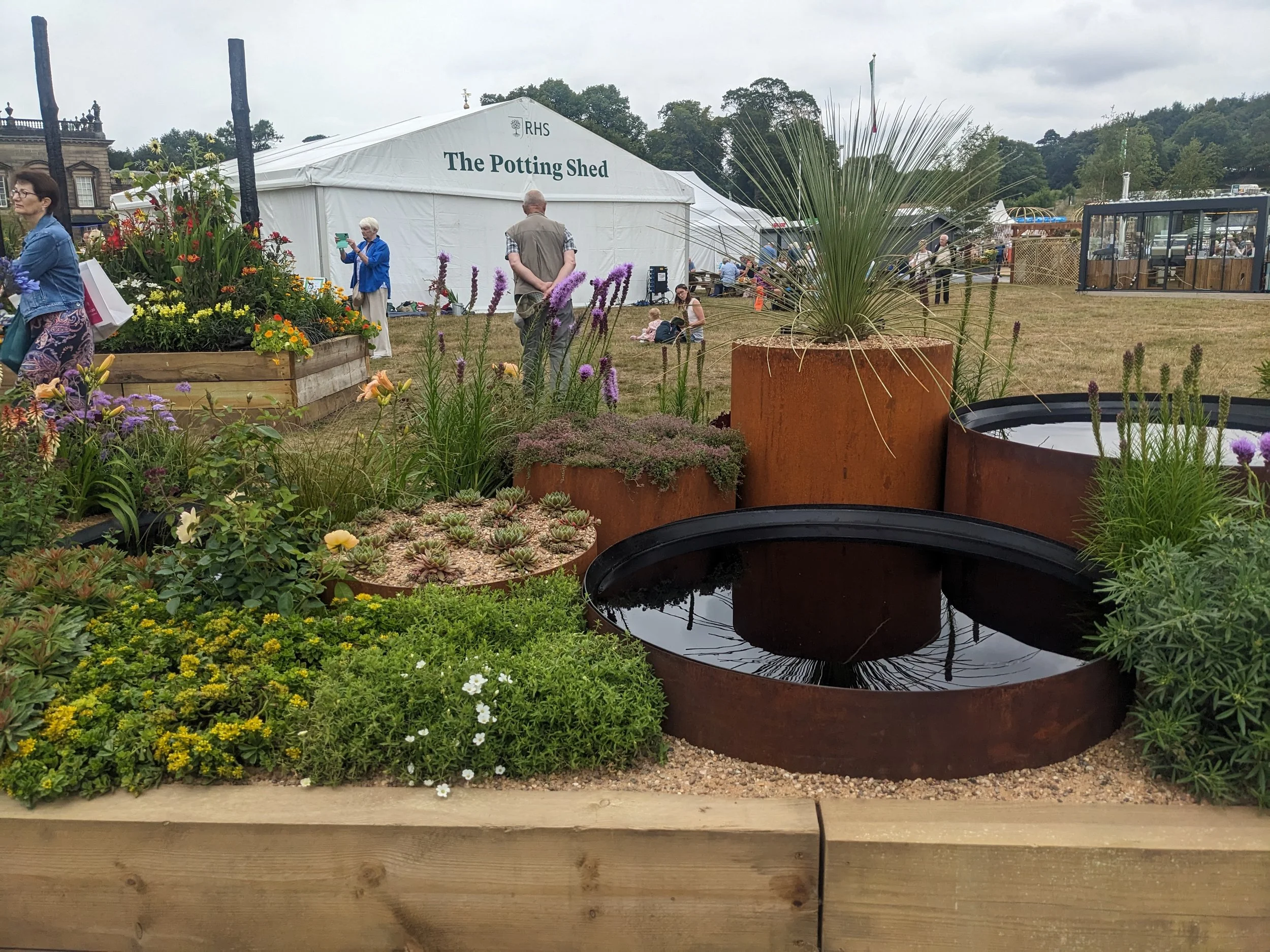 A display of potted plants and flowers at an outdoor garden event, with a large tent labeled 'The Potting Shed' in the background and people walking and sitting nearby.