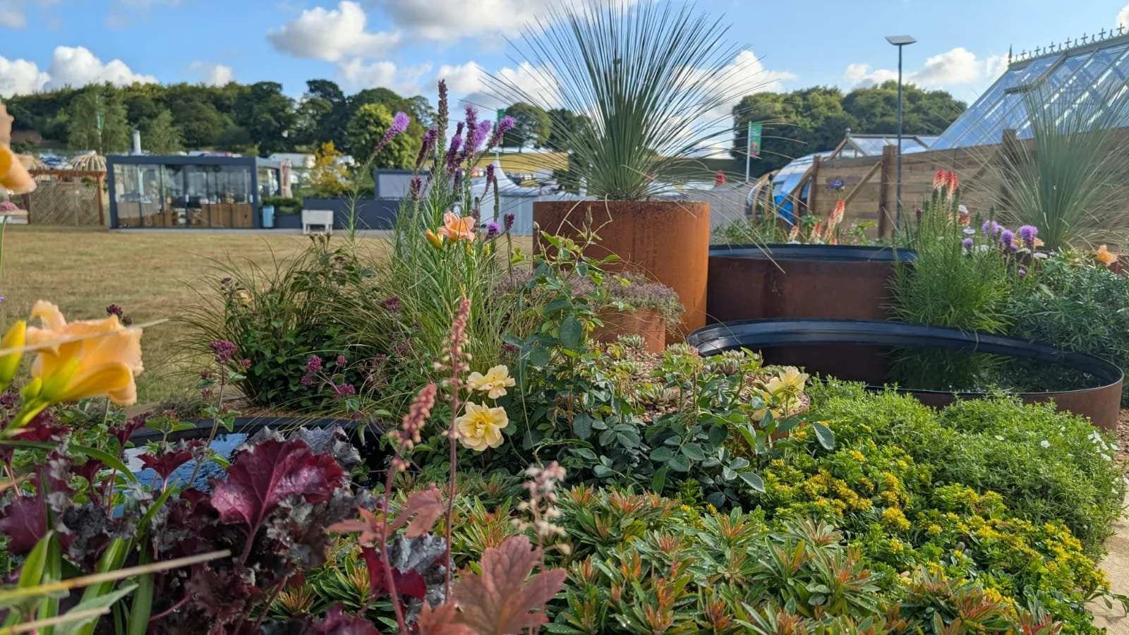 Various potted plants and flowers in an outdoor garden, with greenhouses and a lawn in the background under a partly cloudy sky.