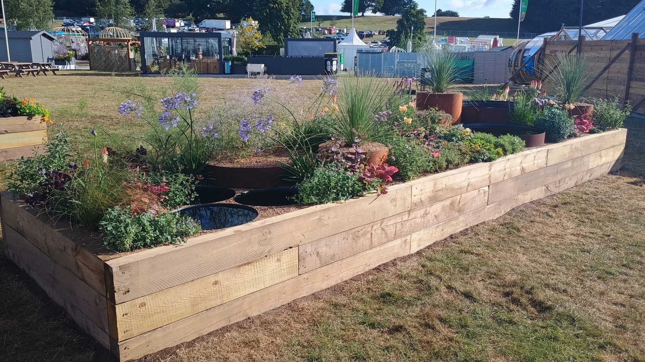 A long wooden raised garden bed filled with various flowering plants and shrubs, situated on a grassy area with a fenced outdoor space and tents in the background.