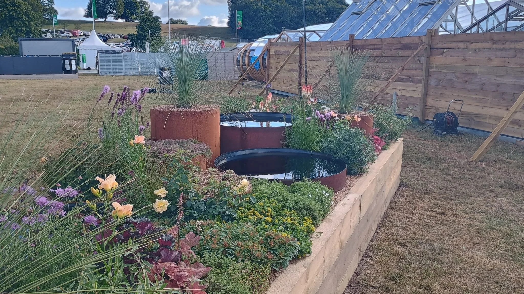 A garden with various flowering plants and tall grasses in large rusty metal containers, enclosed by a wooden fence, with a temporary event setup visible in the background.