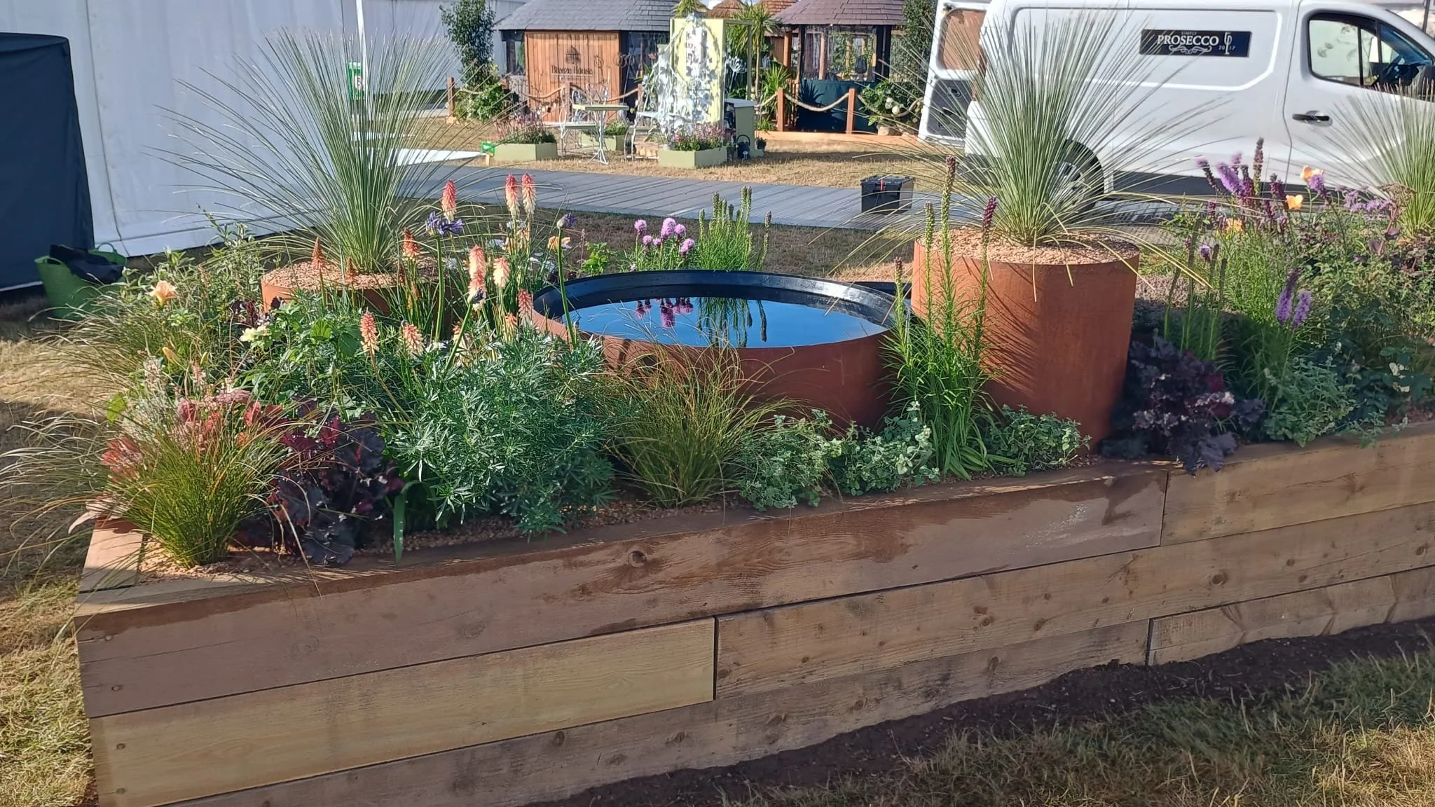 A rectangular wooden planter box filled with various colorful flowers and plants, including tall grasses and purple flowers, with a water feature in the middle. In the background, there are tents and a white van.
