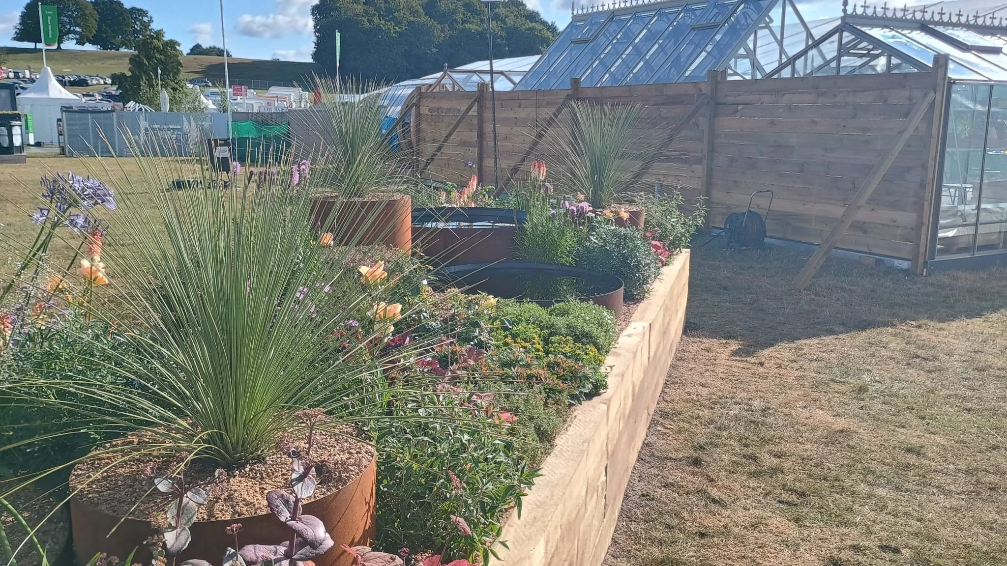 Raised brick garden bed with various flowering plants and tall grasses, beside a wooden fence and greenhouse structures in the background.