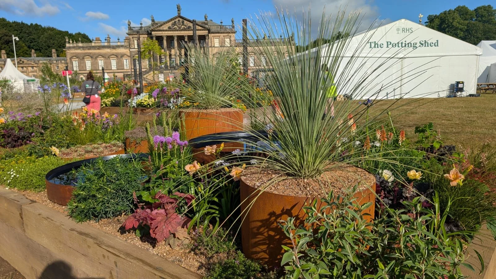 A garden display with potted plants, including tall grasses and blooming flowers, at an outdoor event. A historic building and white tents are visible in the background.