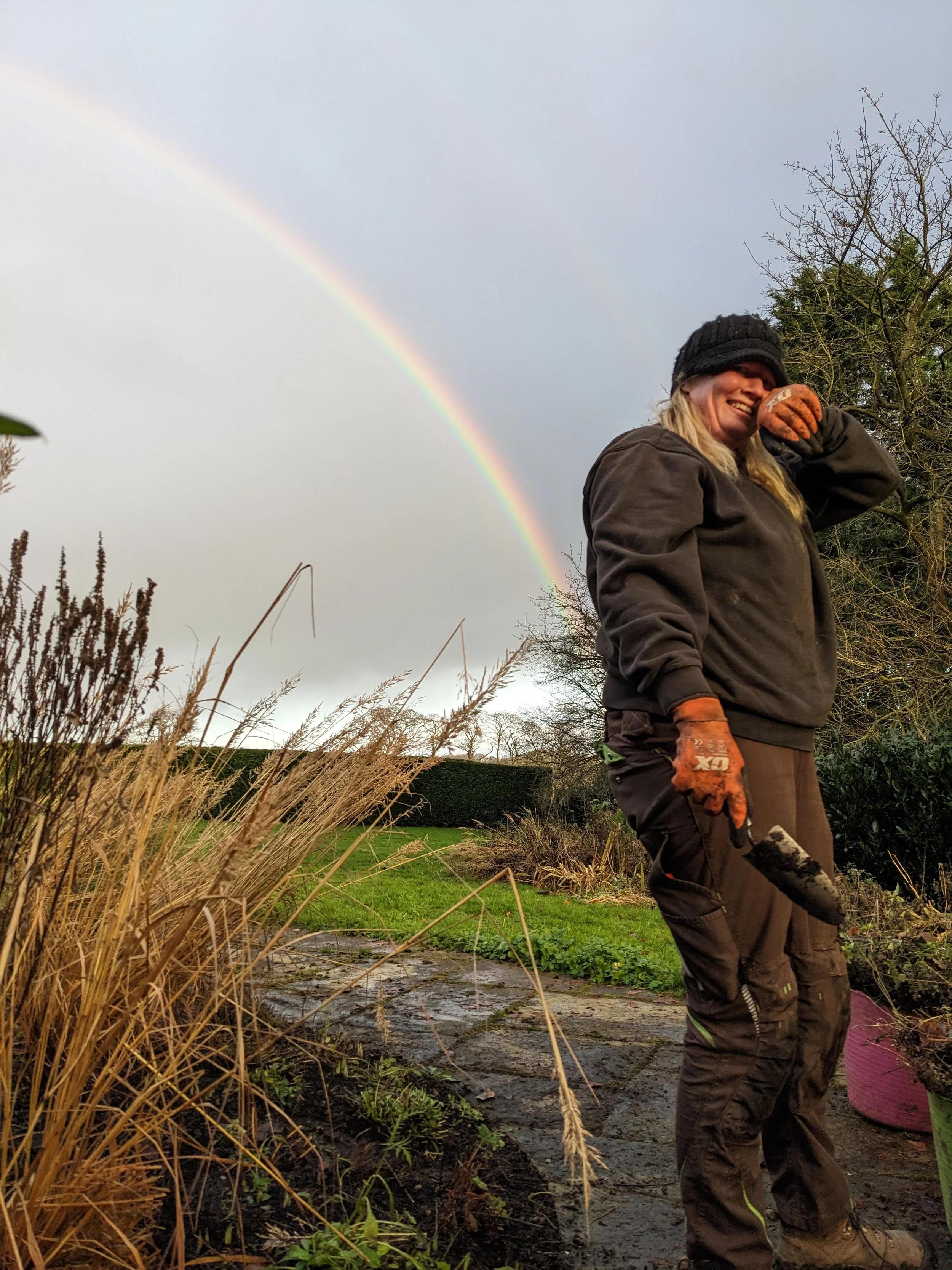 A woman with long brown hair smiling outdoors with trees and greenery in the background.