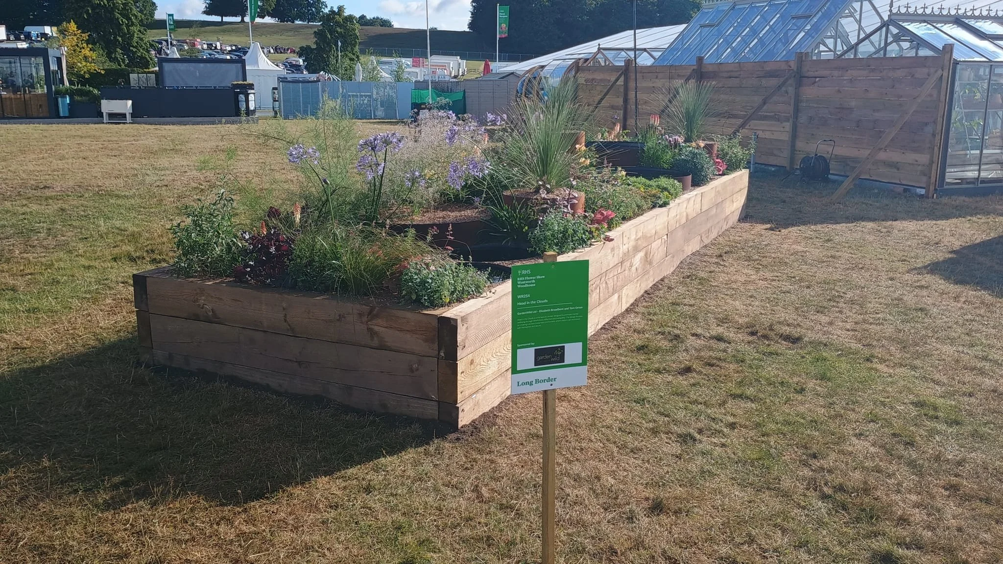 Wooden garden bed with blooming flowers, plants, and greenery on display at an outdoor event, with a sign reading 'Long Border' in front.