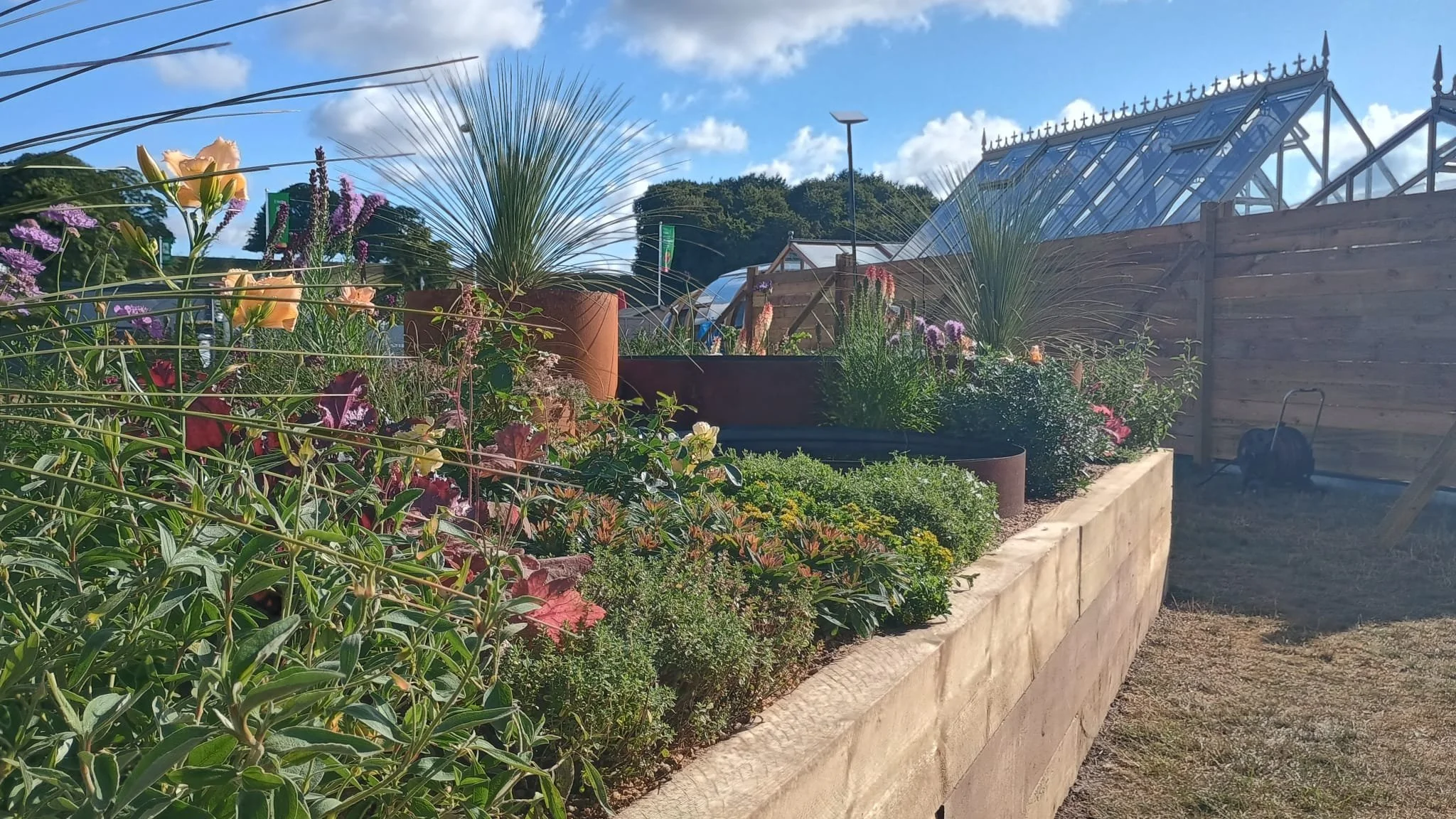 A garden bed filled with colorful flowers and green plants, beside a wooden fence under a partly cloudy sky.