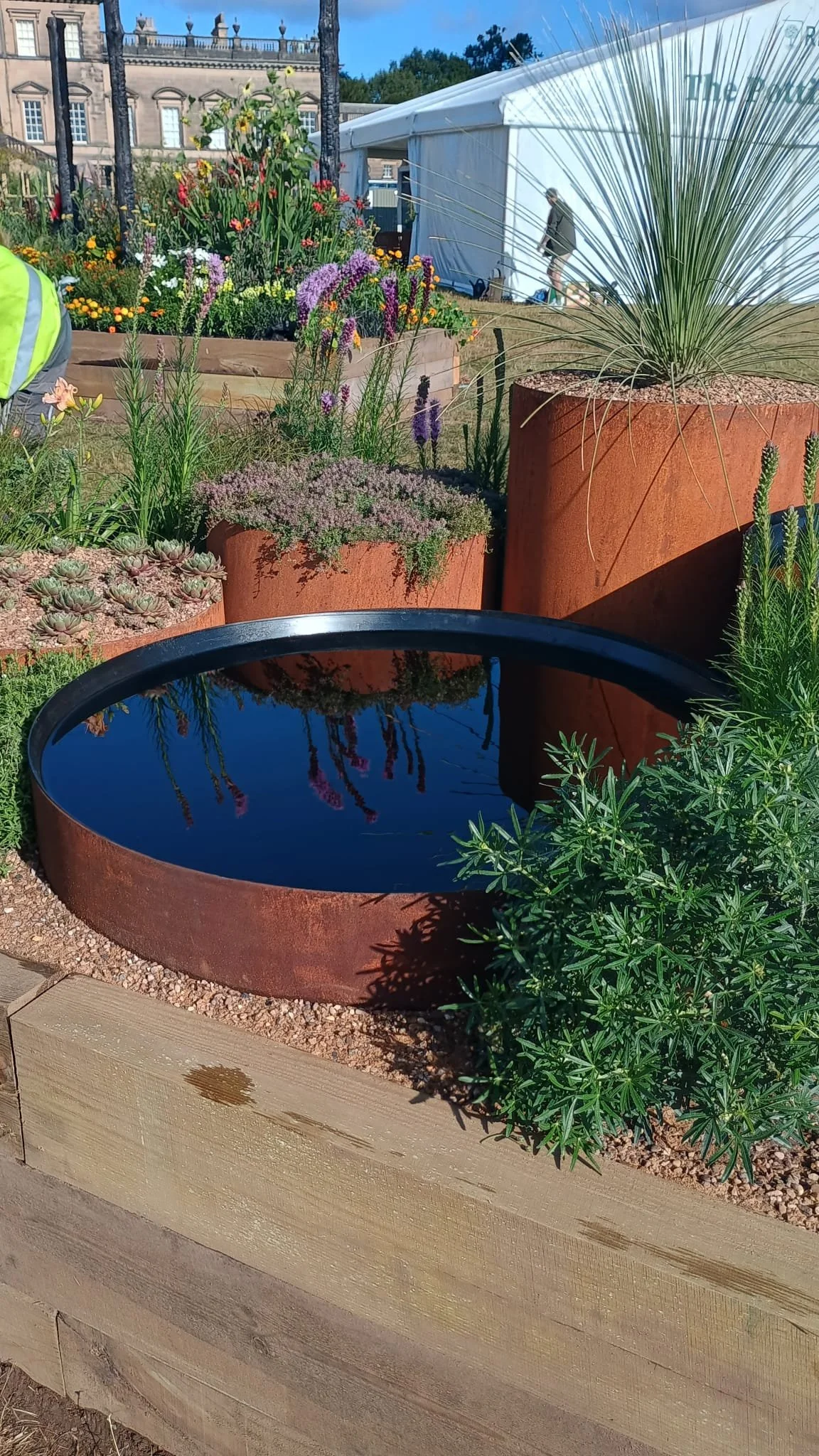 A small round water feature with a black interior, surrounded by rust-colored metal and plants, in a garden with variety of colorful flowers and large planters, with a historic building and white tent in the background.