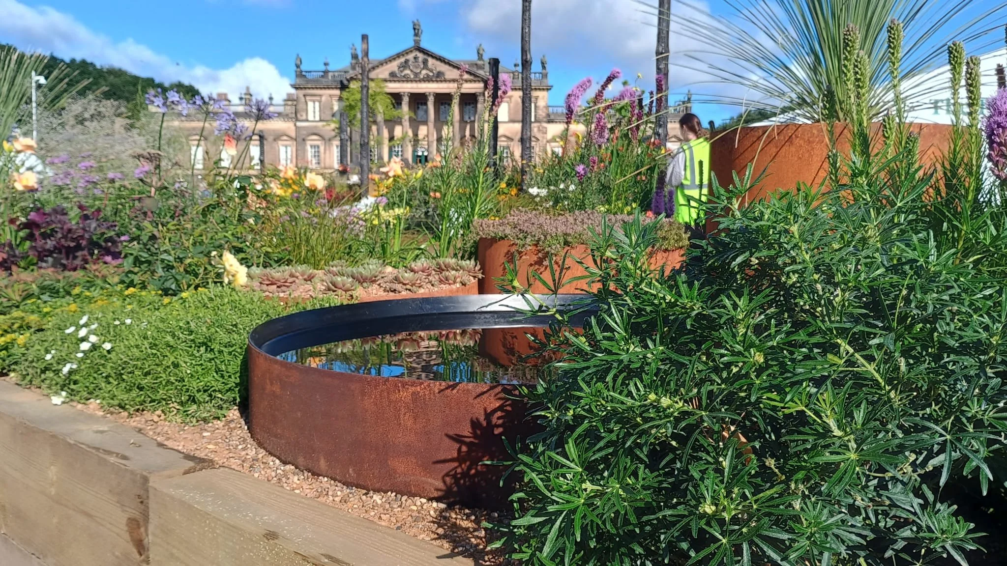 Colorful plants and flowers in a garden with a historic building in the background, and a person wearing a yellow safety vest walking among the plants.