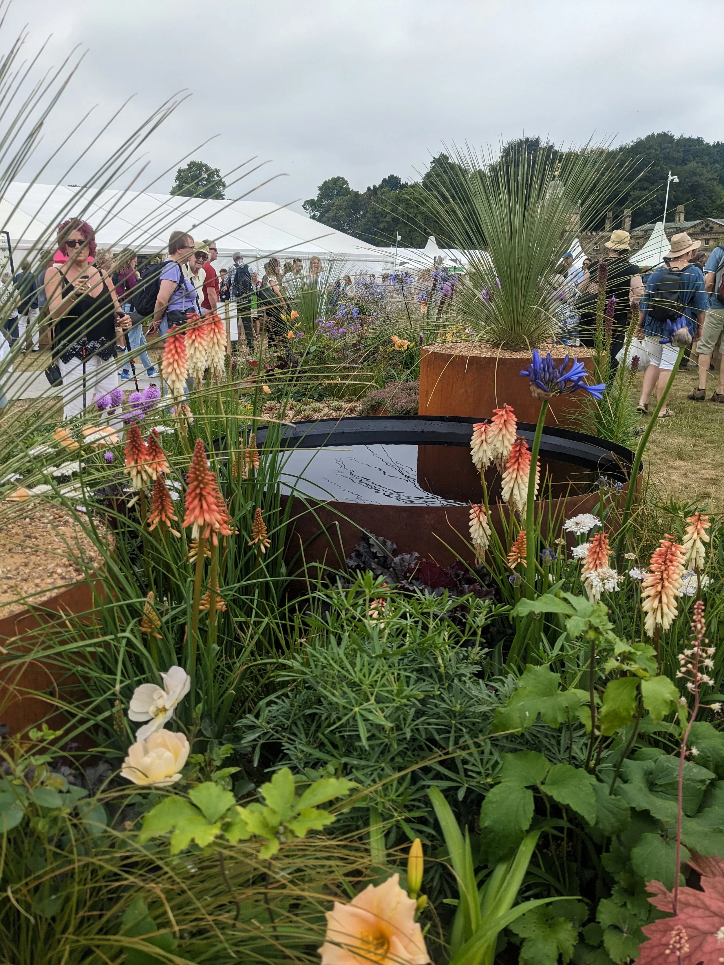 A garden with various colorful flowers and greenery in front of a crowd at an outdoor event with white tents and trees in the background.