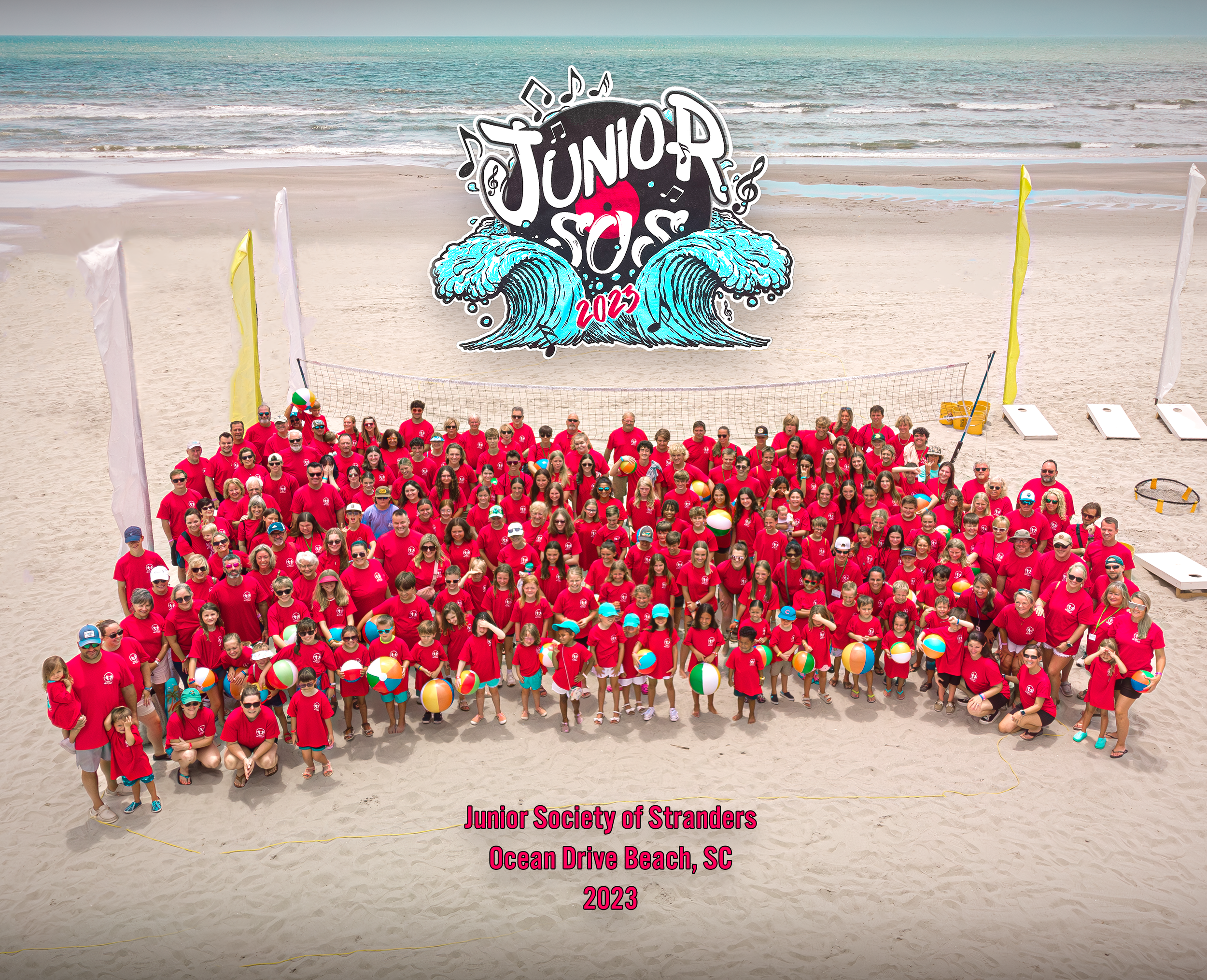 Group of children and adults wearing red shirts on the beach with volleyballs and beach accessories. A large banner with waves and the words 'Junior Beach 2023' is in the background. The text at the bottom reads, 'Junior Society of Stranders, Ocean D
