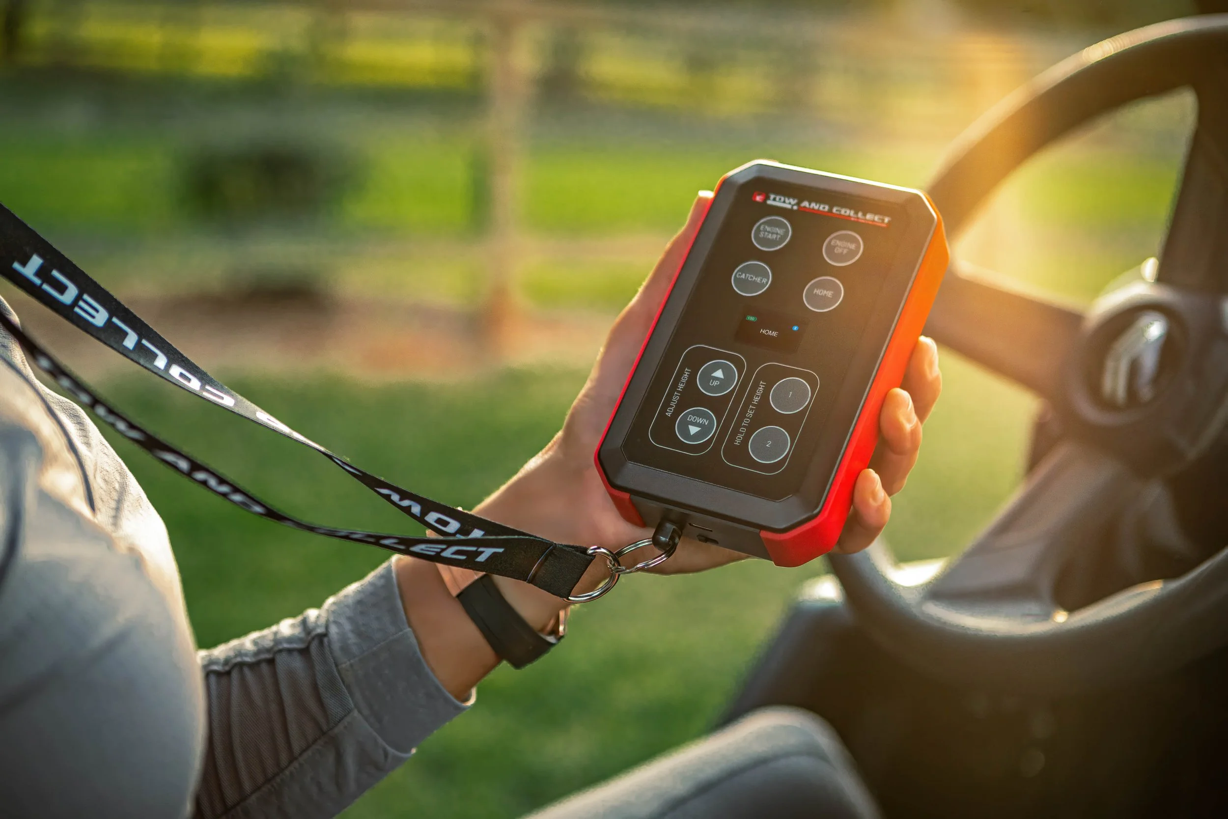 A person holding a remote control device inside a vehicle, with a lanyard around their neck that says 'Action Connect'. The device has buttons and a display screen, and the vehicle's steering wheel is visible to the right. The background shows a gras