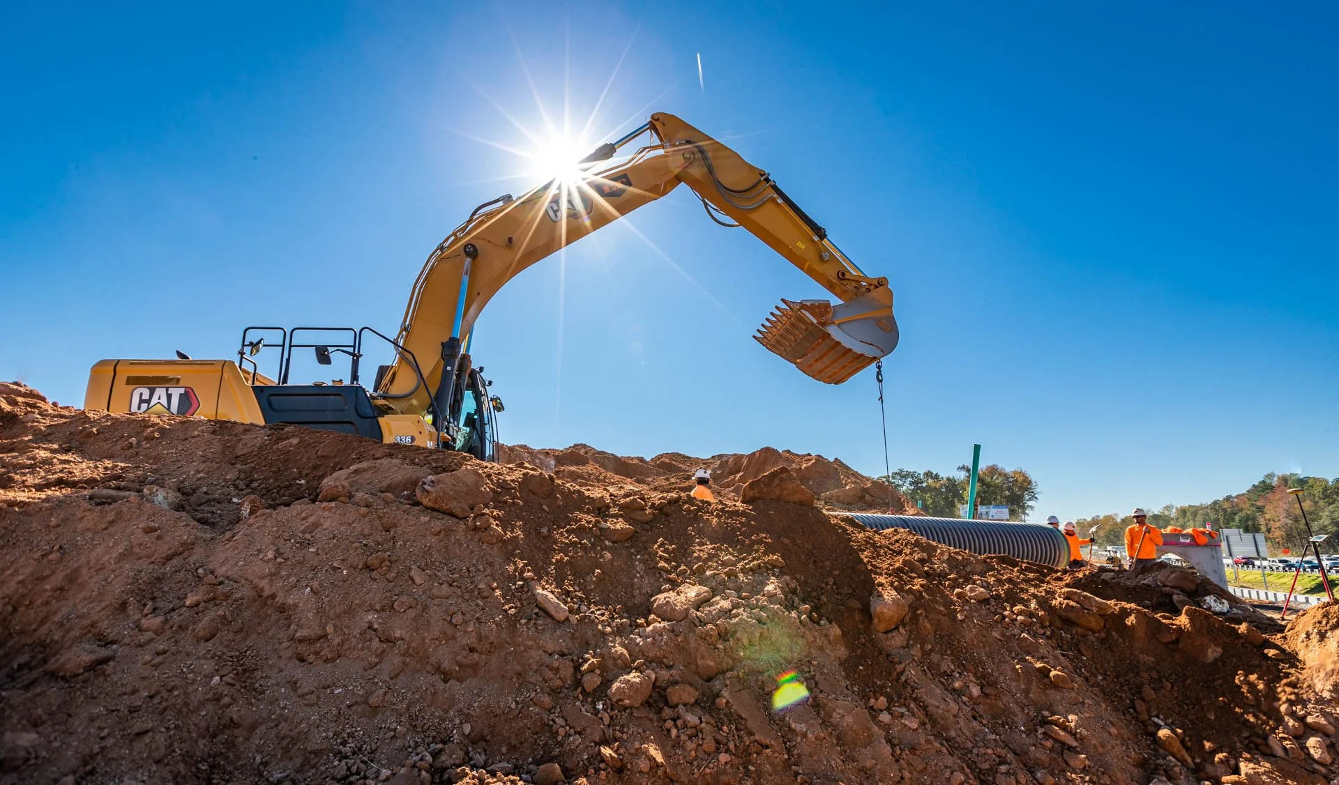 A construction site with an excavator digging into the dirt, sunlight shining behind the machine, and workers in orange safety shirts and white helmets in the background.