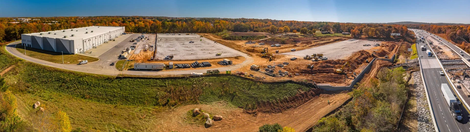 Construction site next to a large warehouse and a highway, with ongoing earthwork and machinery.