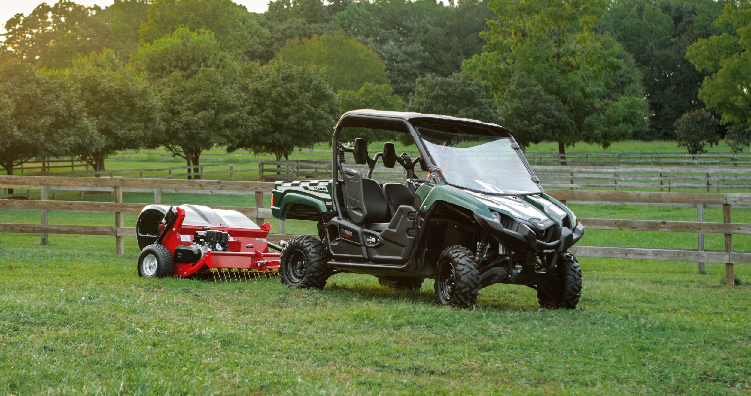A green utility vehicle with a black roof and side doors, parked on a grassy field. Attached to it is a red mower with black and white wheels, used for cutting grass. In the background, there are wooden fences and numerous trees, indicating a rural o